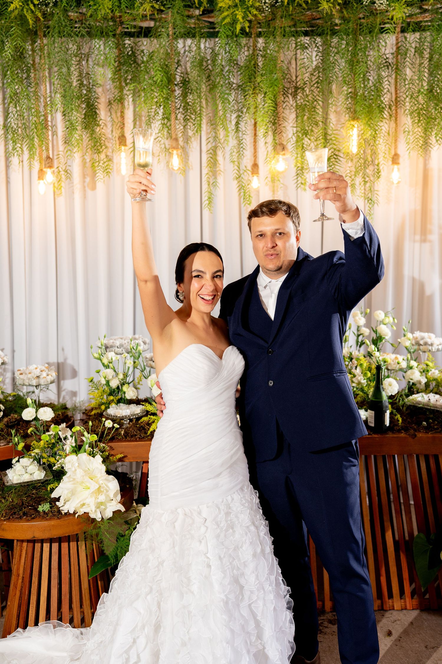 Bride and groom raising champagne glasses in toast at elegant wedding reception with floral decor and hanging lights.