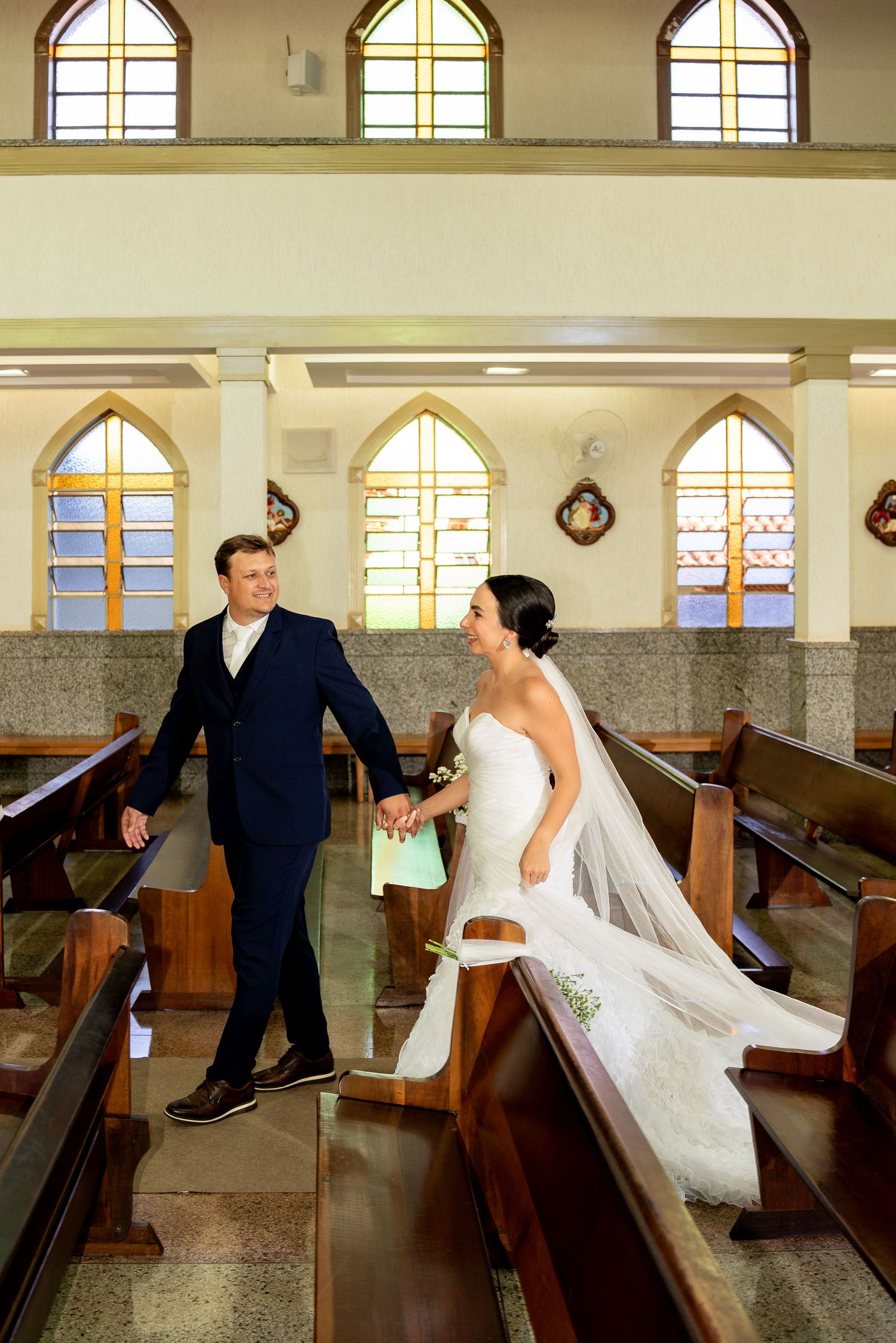 Bride and groom walk hand in hand down church aisle in elegant wedding attire, surrounded by wooden pews and stained glass windows.