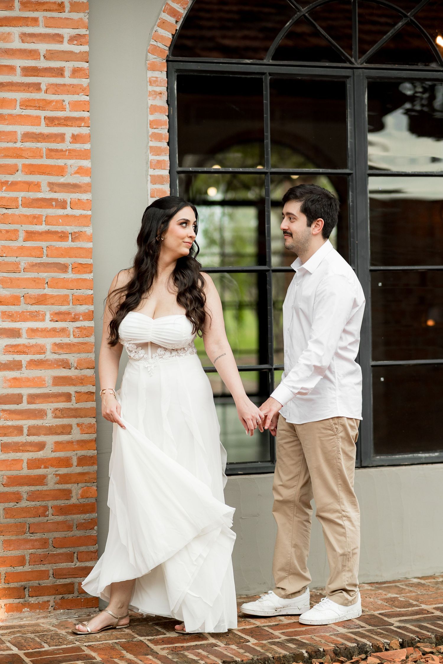 A bride in a white gown and groom in white shirt hold hands outdoors near a brick wall and large window.