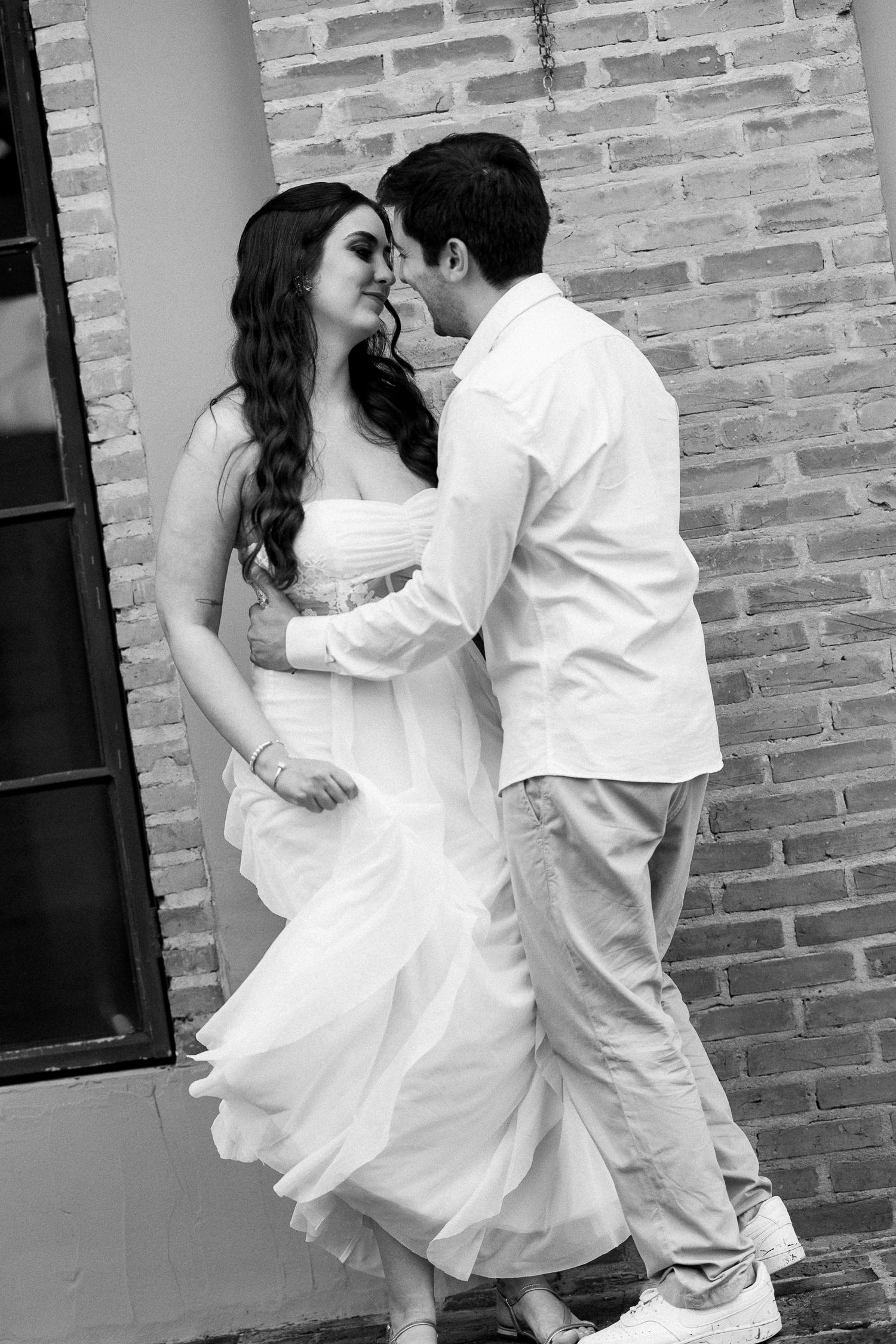 Black and white photo of a couple in wedding attire sharing a romantic moment against a brick wall.