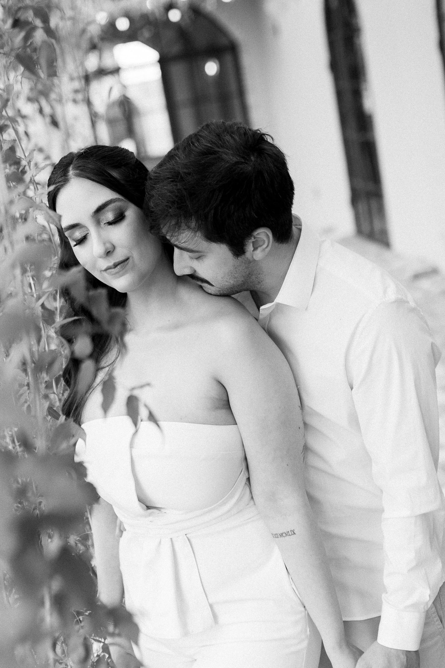 Black and white photo of a romantic couple embracing near lush greenery, woman in strapless white dress, man in white shirt.