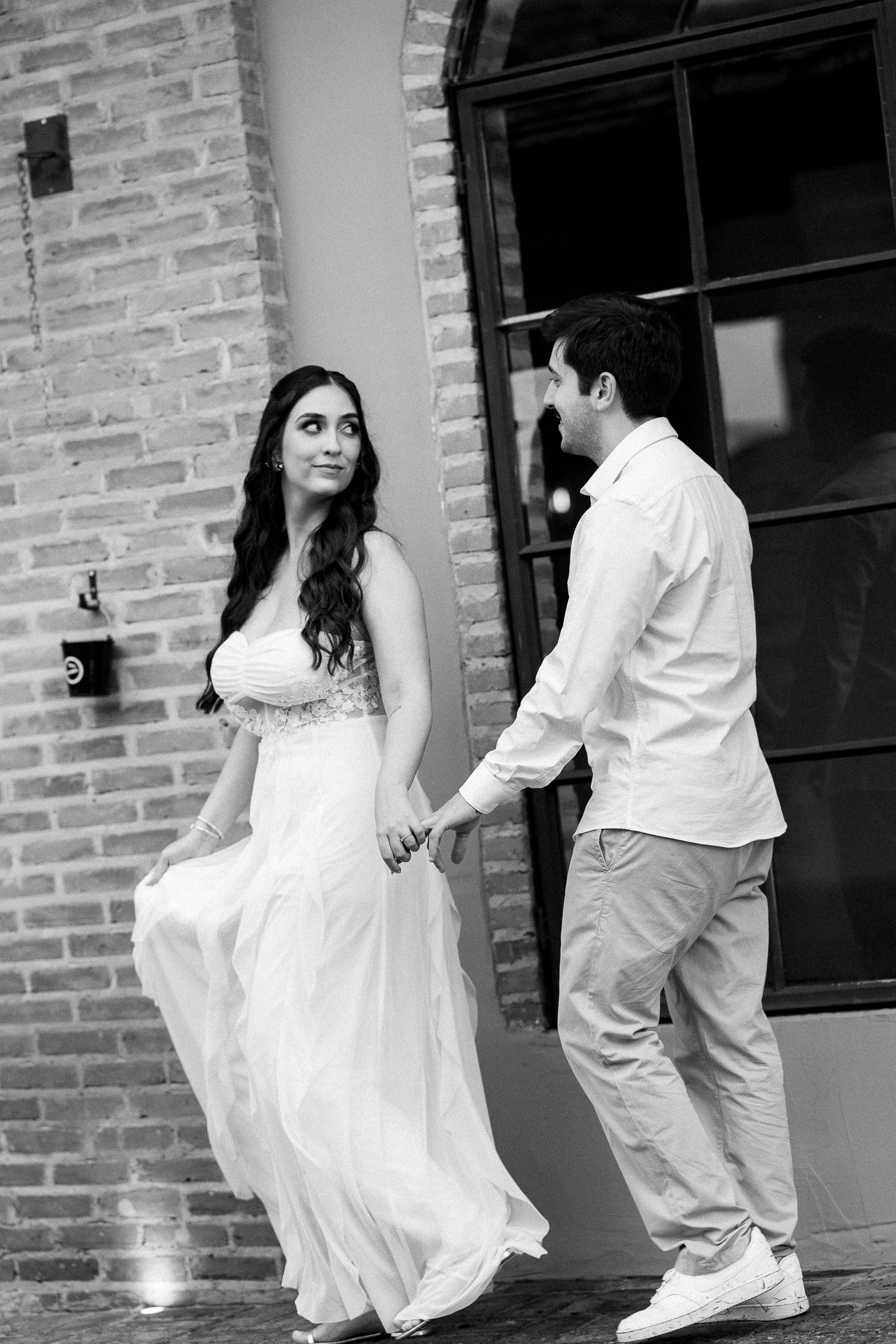 Black and white photo of a couple holding hands, woman in flowing white wedding dress, man in casual attire, brick building backdrop.