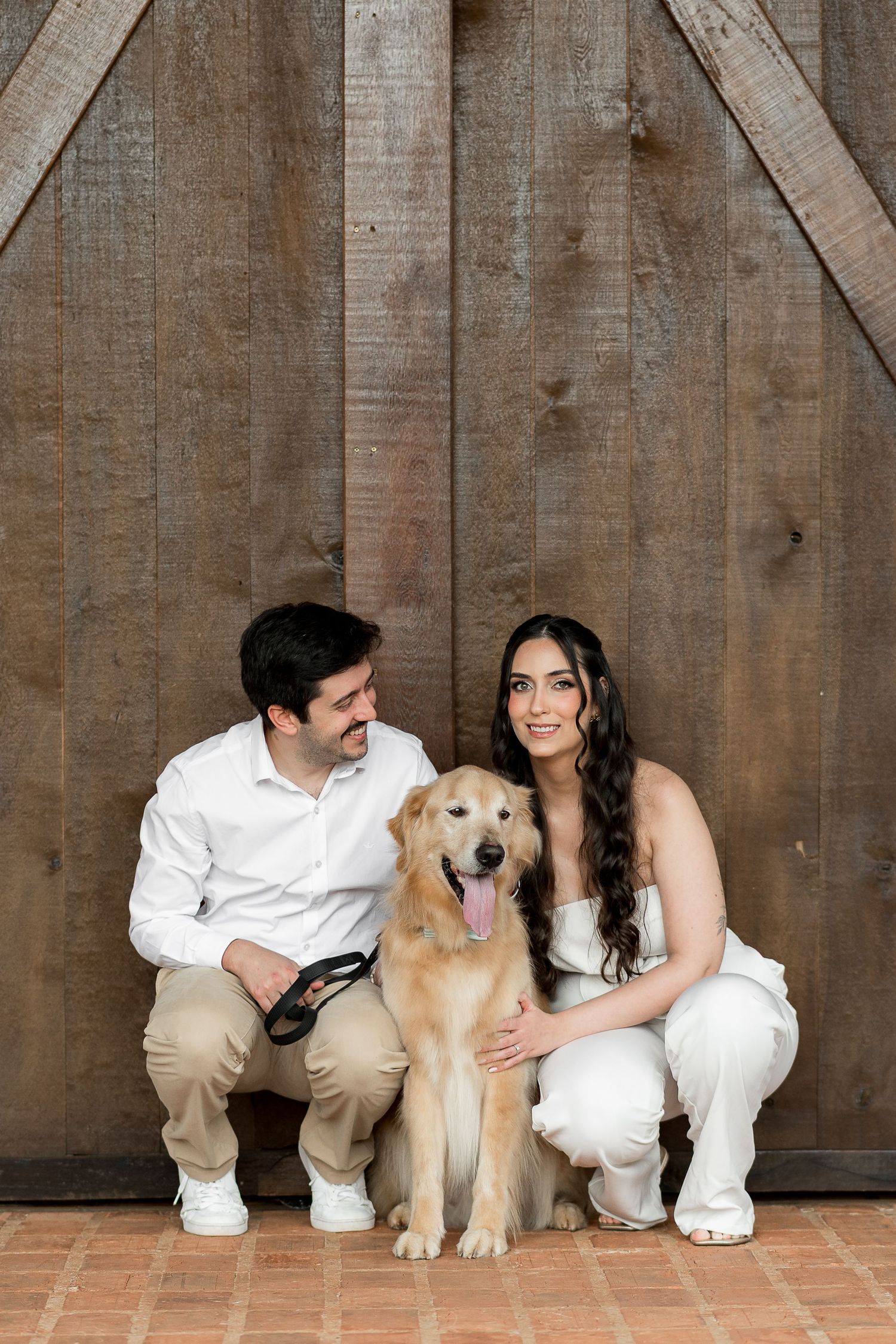 Couple in white outfits crouching with a golden retriever in front of rustic wooden barn doors.