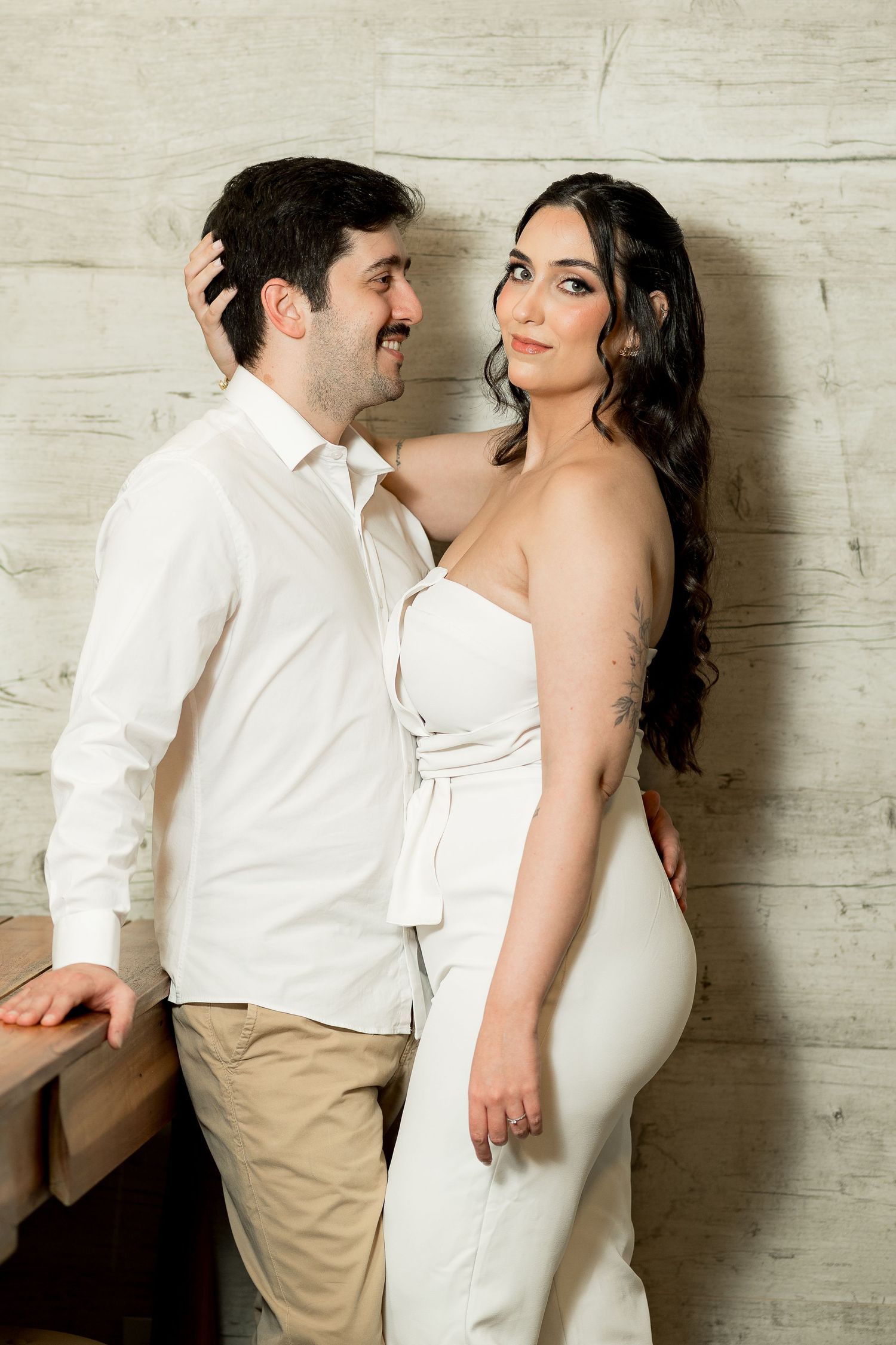 A couple in white outfits poses romantically against a rustic wooden wall, the woman in a strapless jumpsuit smiling at the camera.