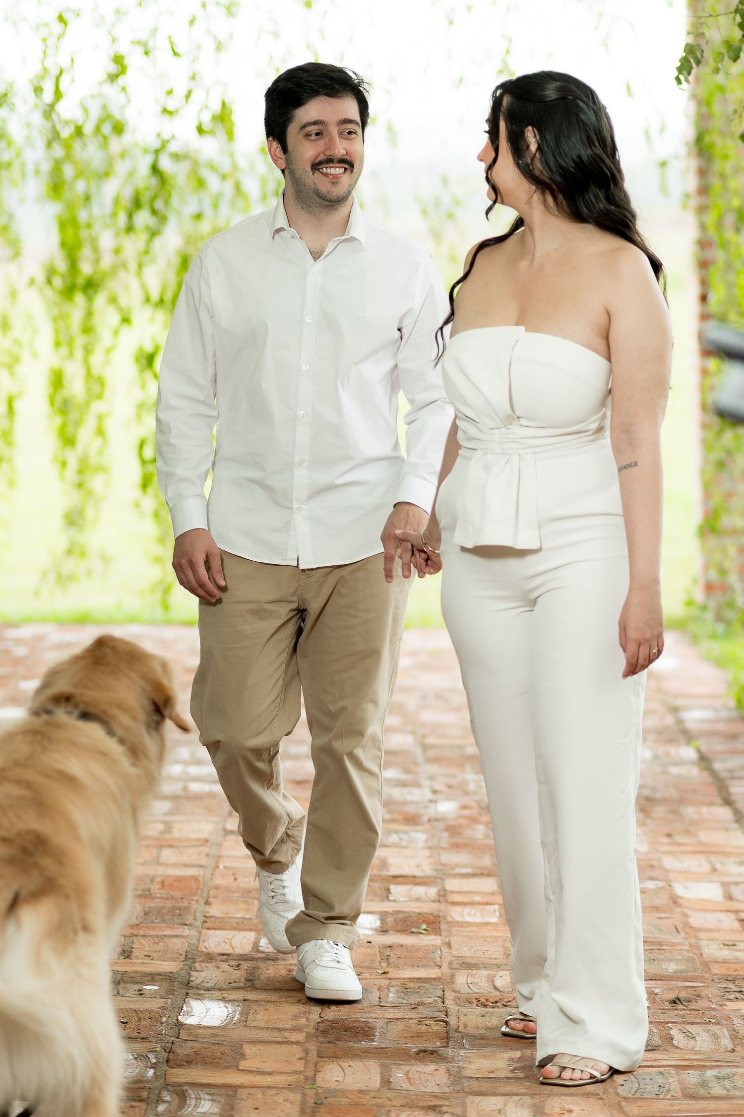 Couple holding hands walking on brick path with golden retriever, man in white shirt and woman in white outfit.