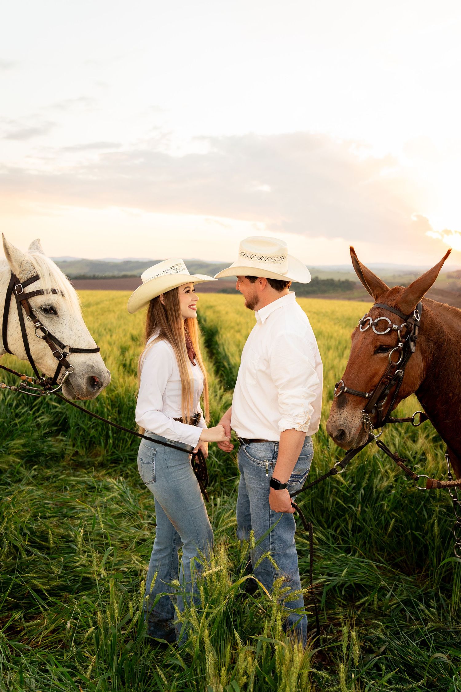 Couple in cowboy hats holding hands in a green field at sunset with a white and brown horse behind them.