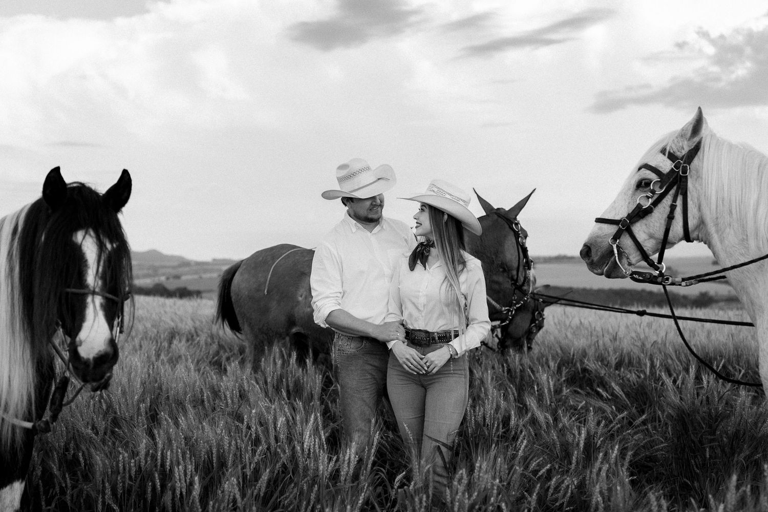 Black and white photo of a couple in cowboy hats standing romantically among horses in a grassy field.