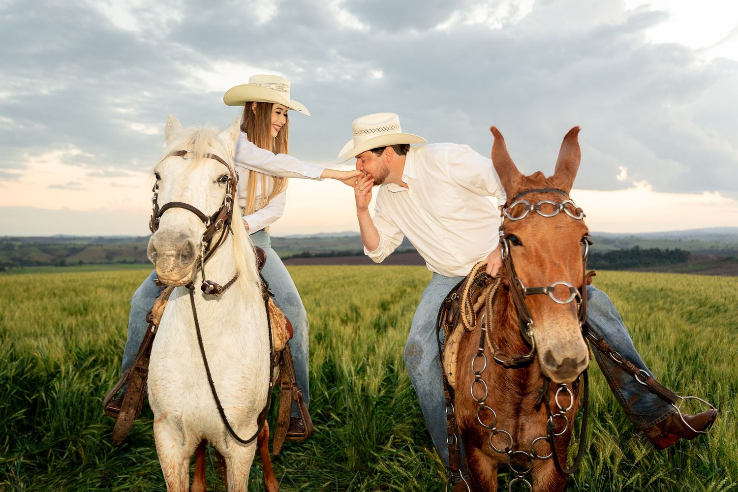 A couple in cowboy hats share a romantic kiss while riding horses in a lush green field.