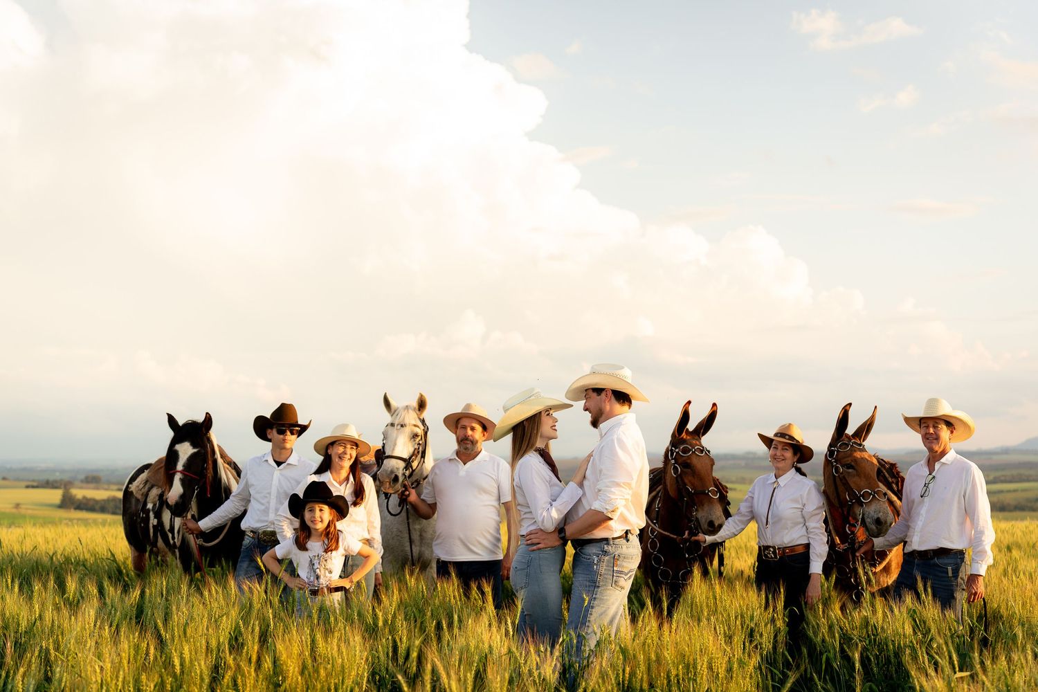 A group of cowboys and cowgirls in white shirts and straw hats stand with horses in a lush green field.