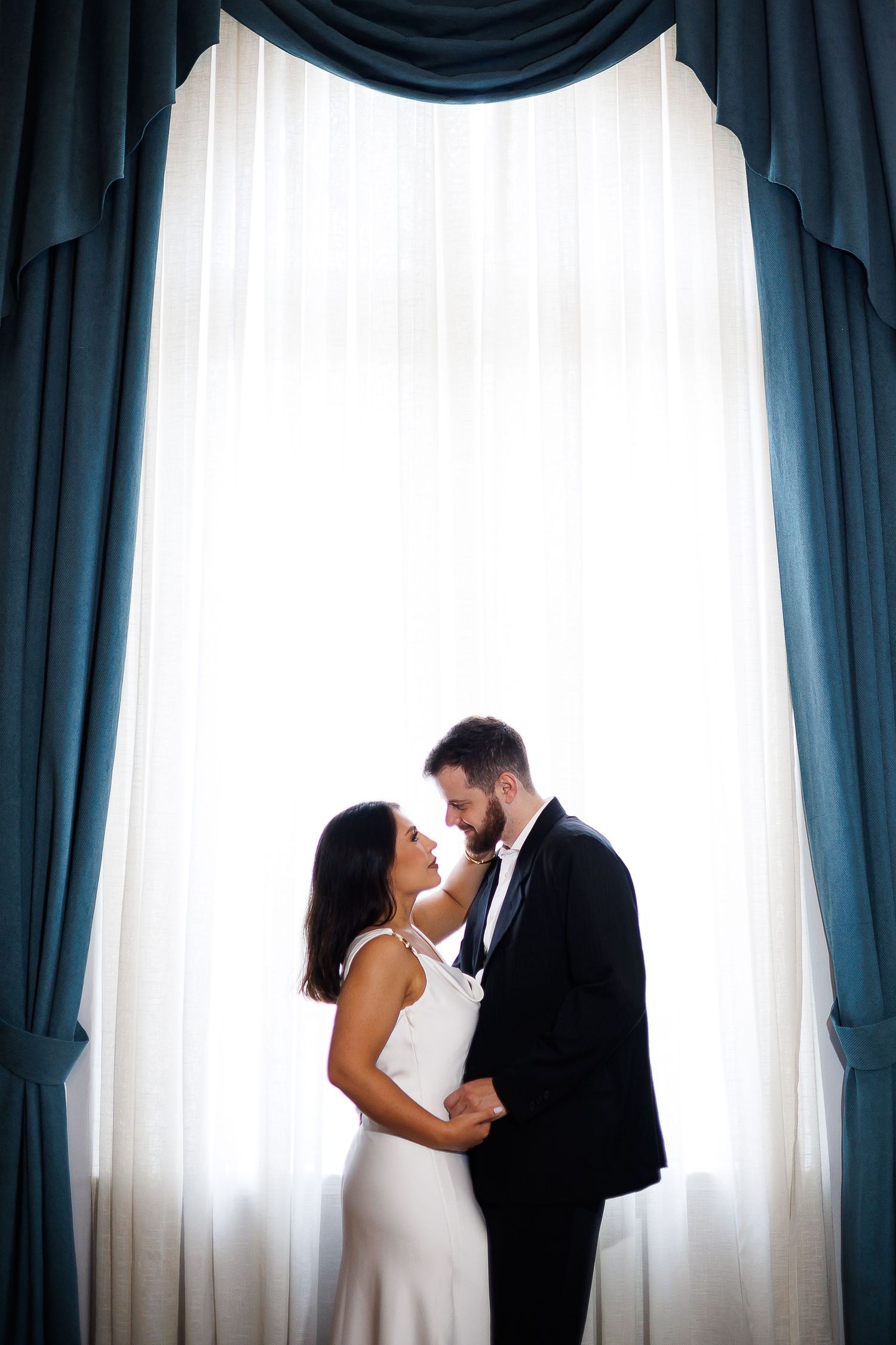 Bride and groom share a romantic moment in front of elegant teal drapes and a bright window.