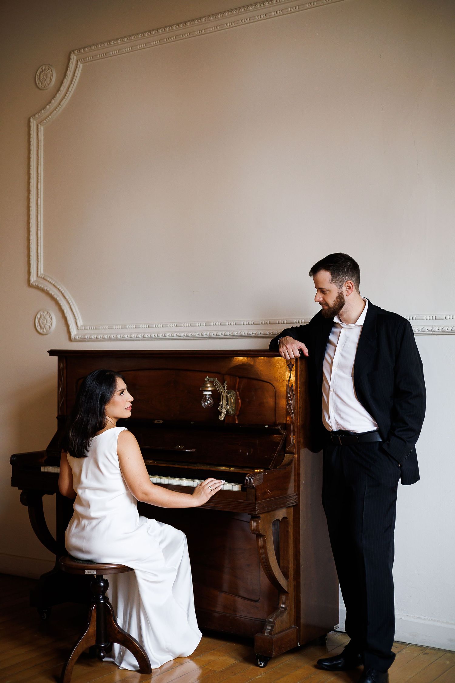 Woman in white dress plays upright piano while man in black suit leans on it, elegant room setting.