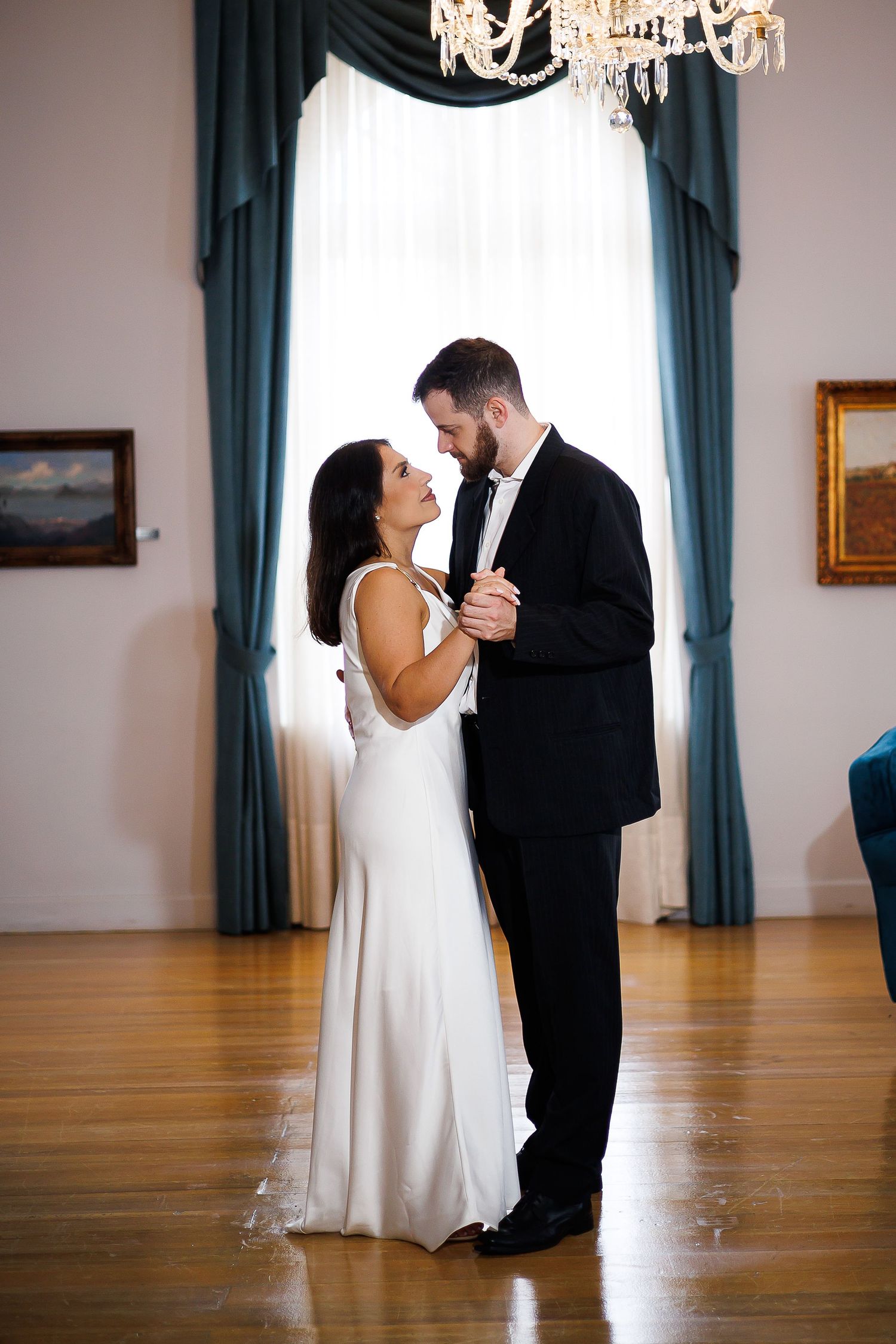 Bride and groom share first dance in elegant ballroom with chandelier and teal drapes.