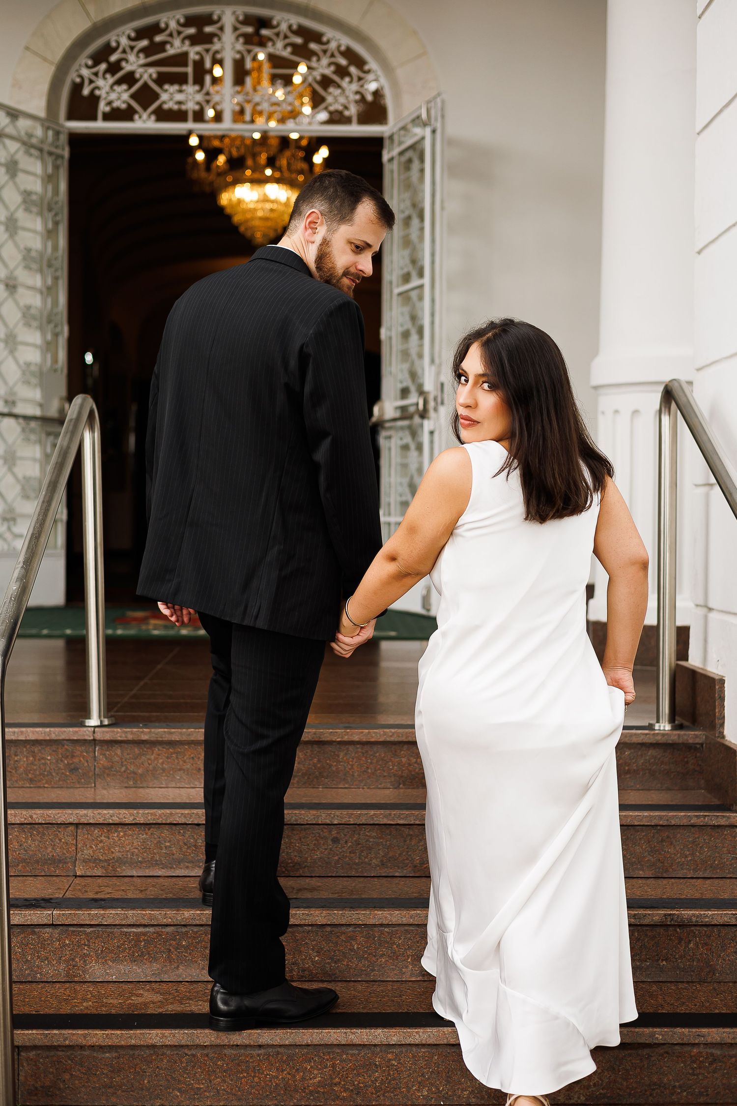 Couple in formal attire holding hands on elegant staircase, woman in white gown looking back, man in black suit.