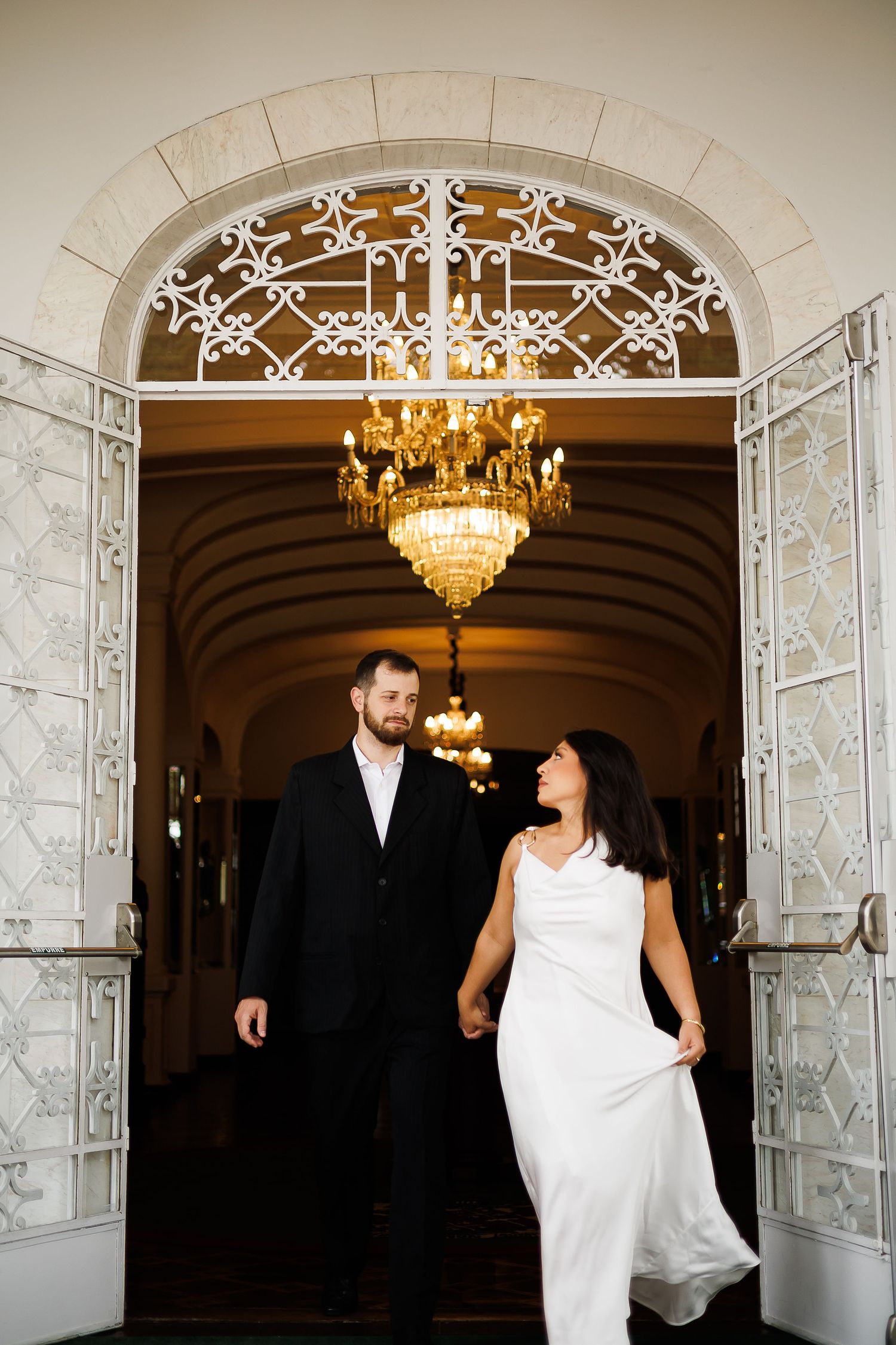 Bride and groom hold hands walking through ornate white doors beneath a golden chandelier in an elegant venue.