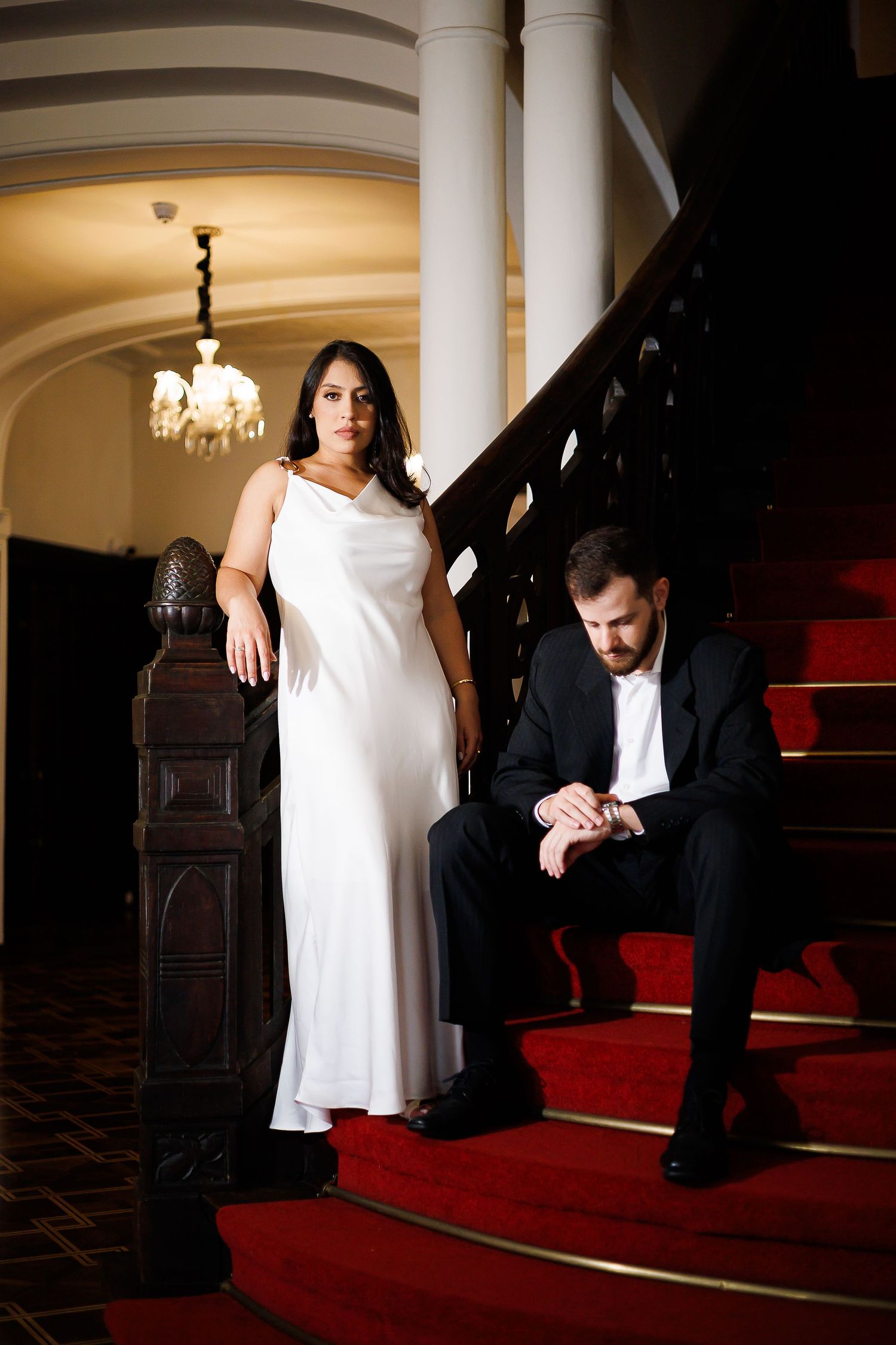 Elegant couple on red-carpeted stairs, woman in white gown standing, man in black suit seated, in a grand interior.
