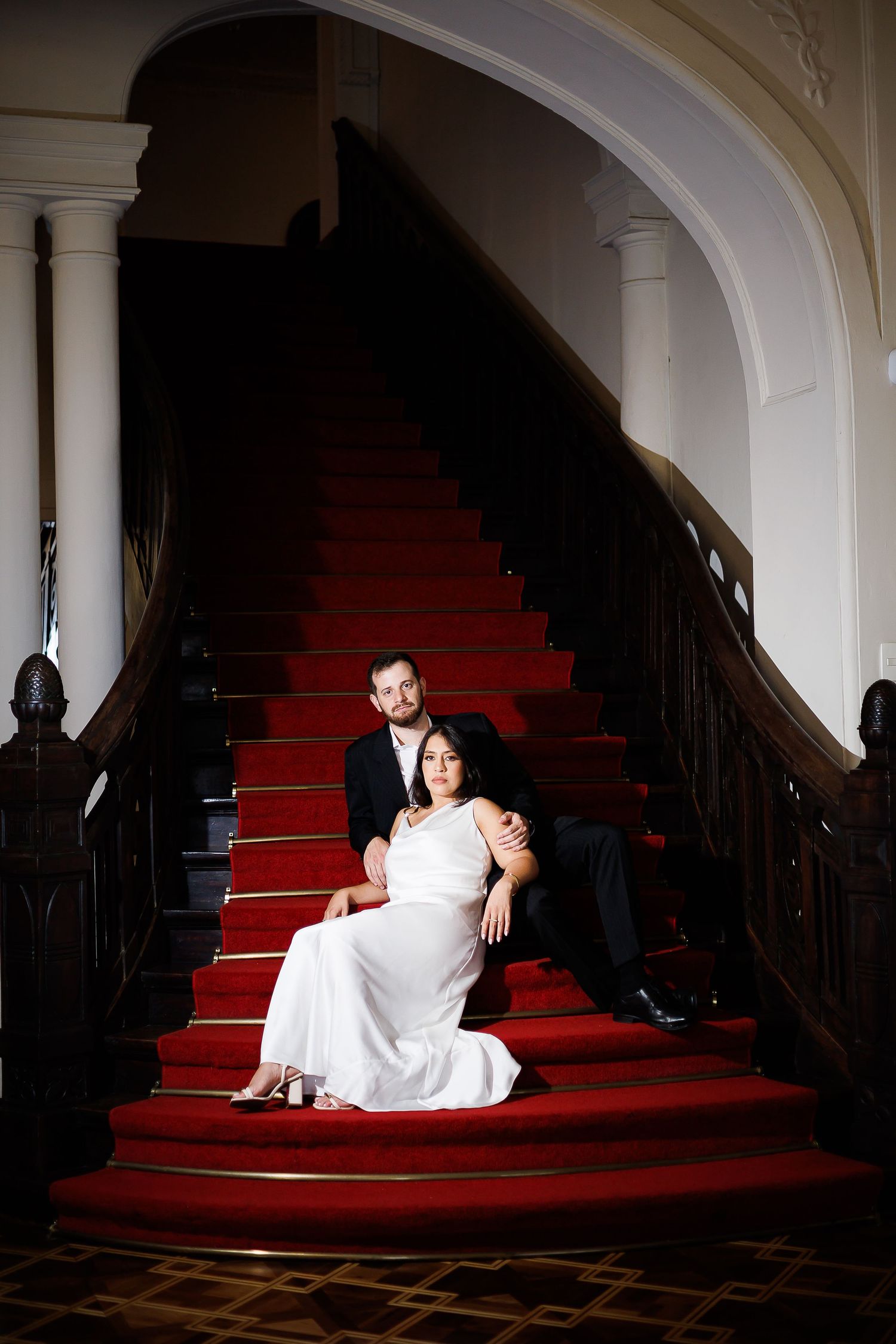 Elegant couple in wedding attire posing on red-carpeted grand staircase in a classic venue.