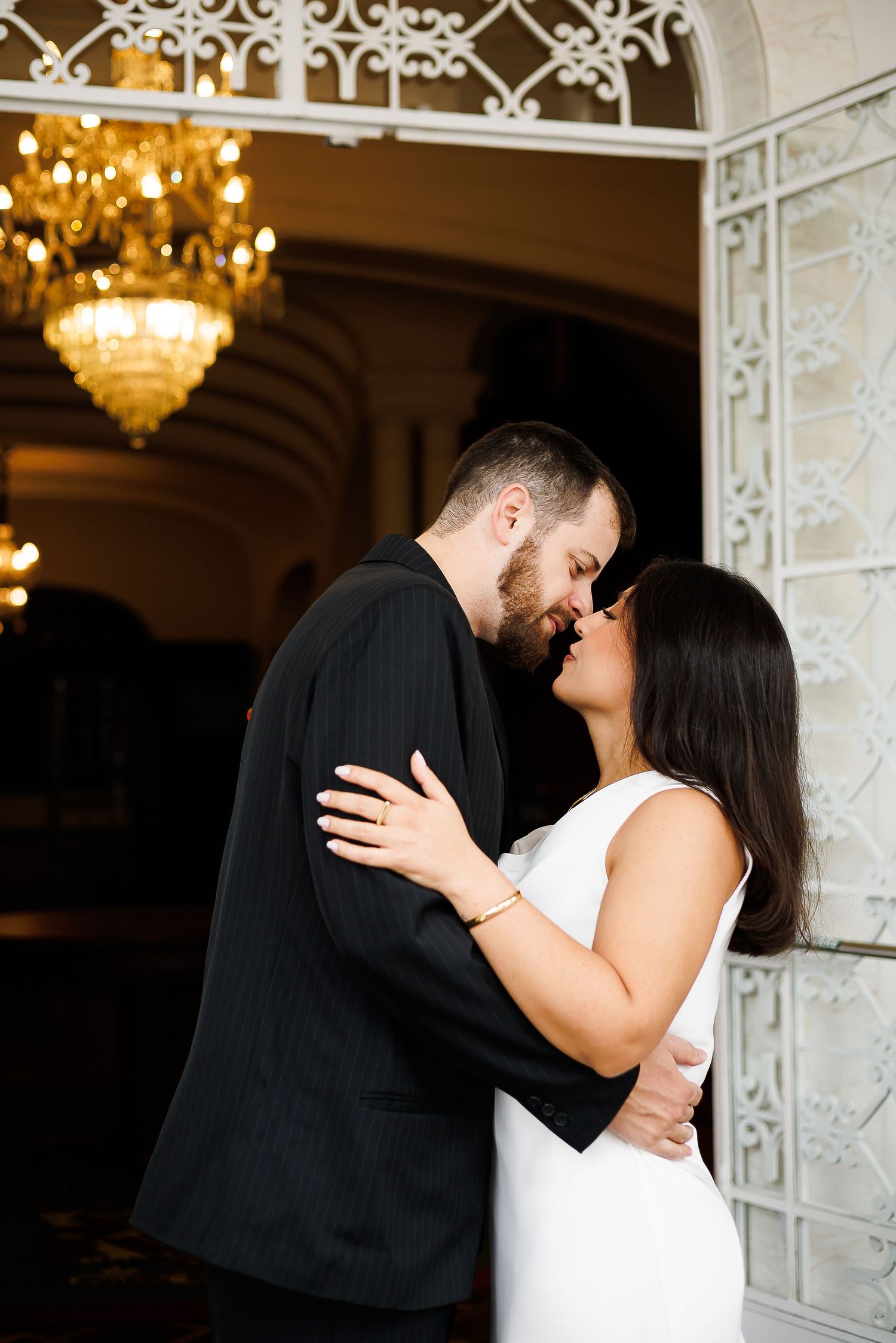 Bride and groom sharing a romantic kiss in an elegant venue with a chandelier, wearing white dress and black suit.