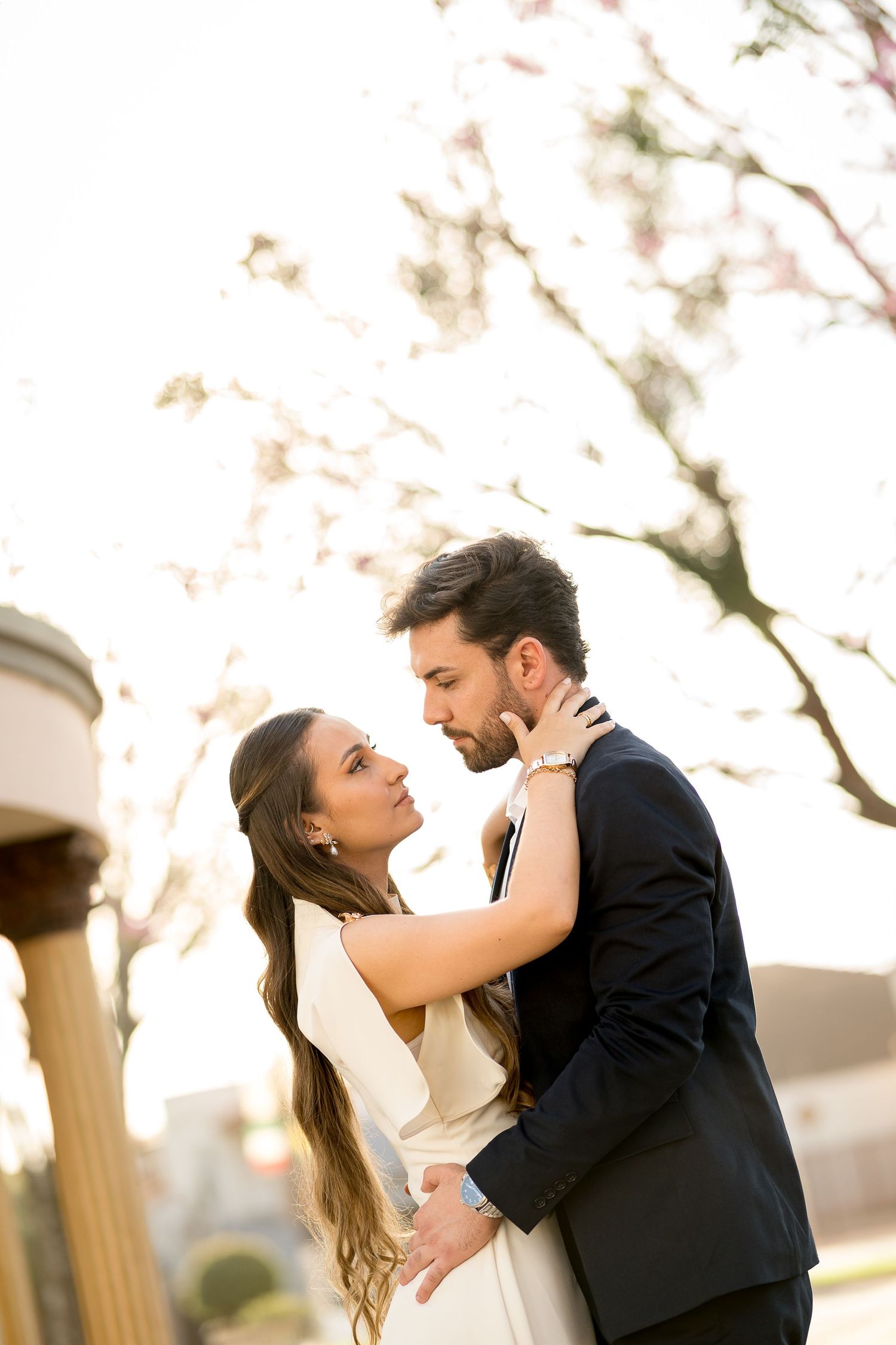 A couple in elegant attire sharing a romantic embrace outdoors, with soft sunlight and blooming trees in the background.