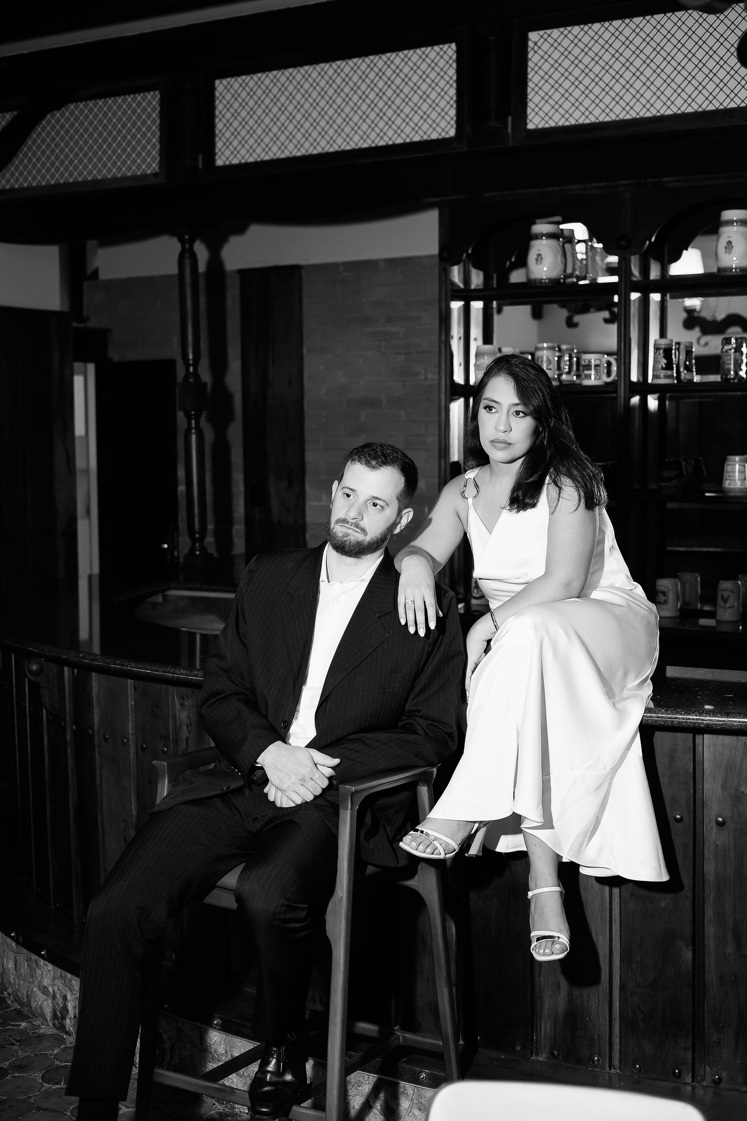 Black and white photo of a couple posing in a bar, man in suit seated, woman in white dress beside him.