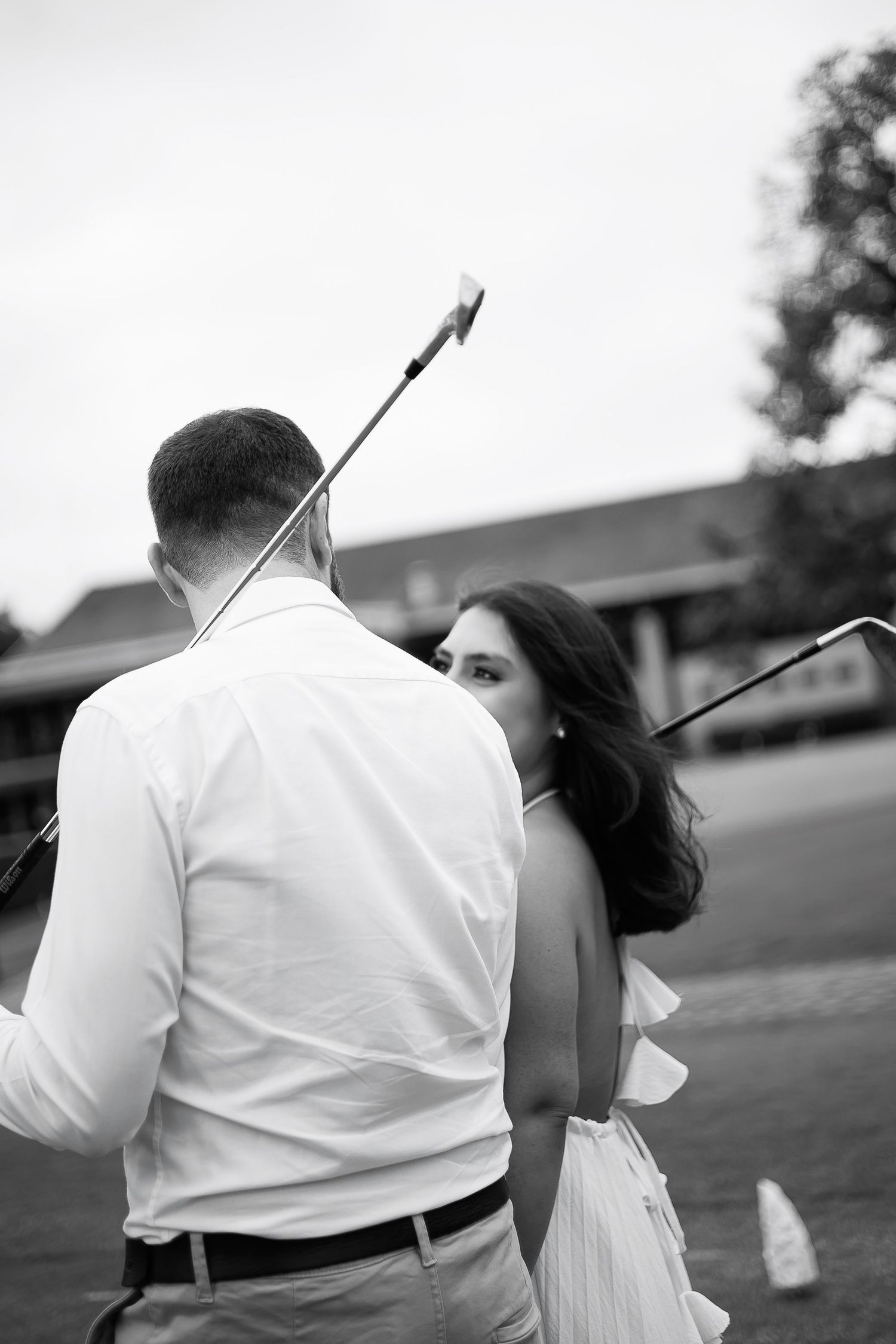 Black and white photo of a couple holding golf clubs on a golf course, woman in backless dress.