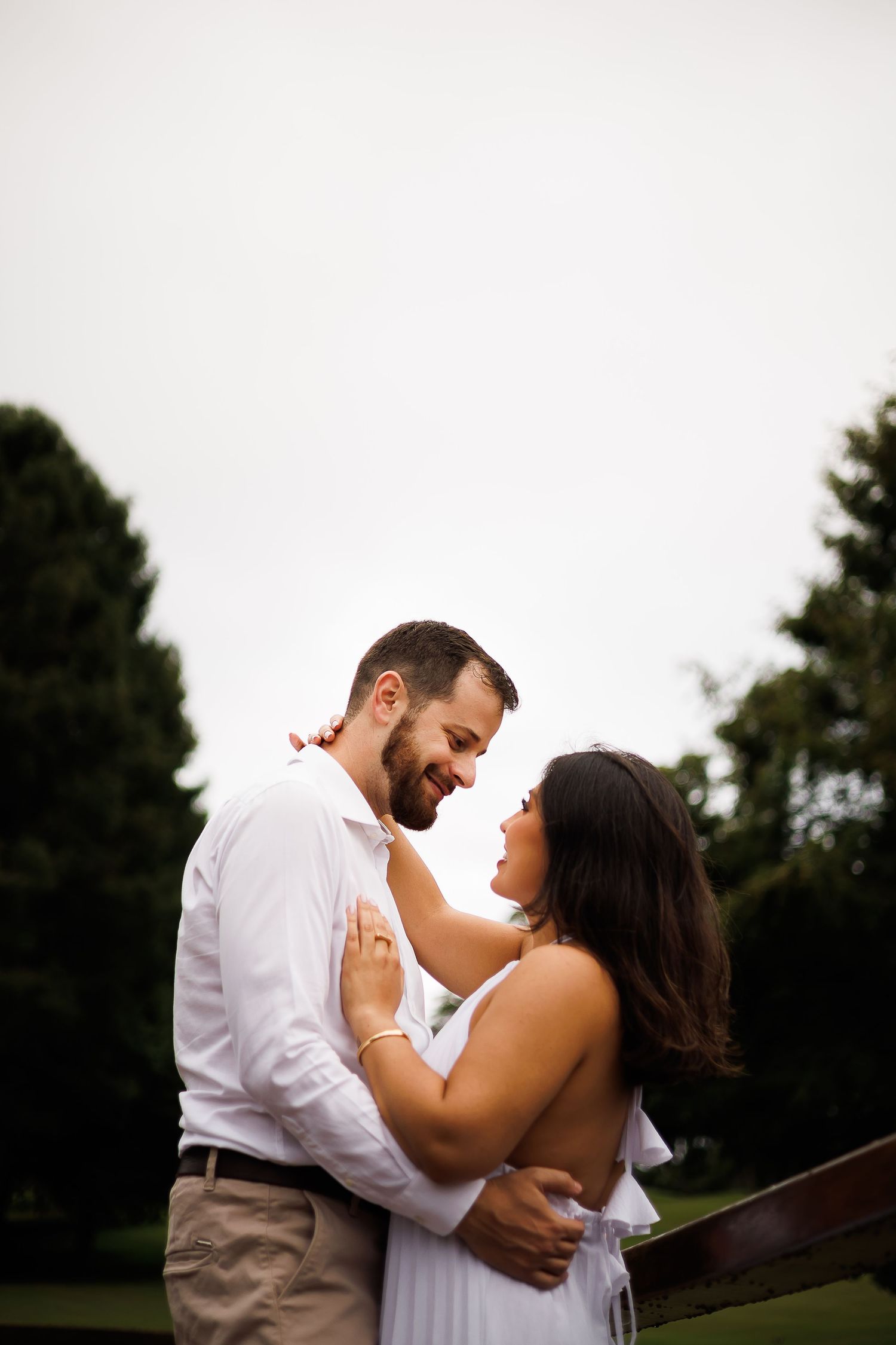 A couple in white attire embraces outdoors, smiling at each other with lush green trees in the background.