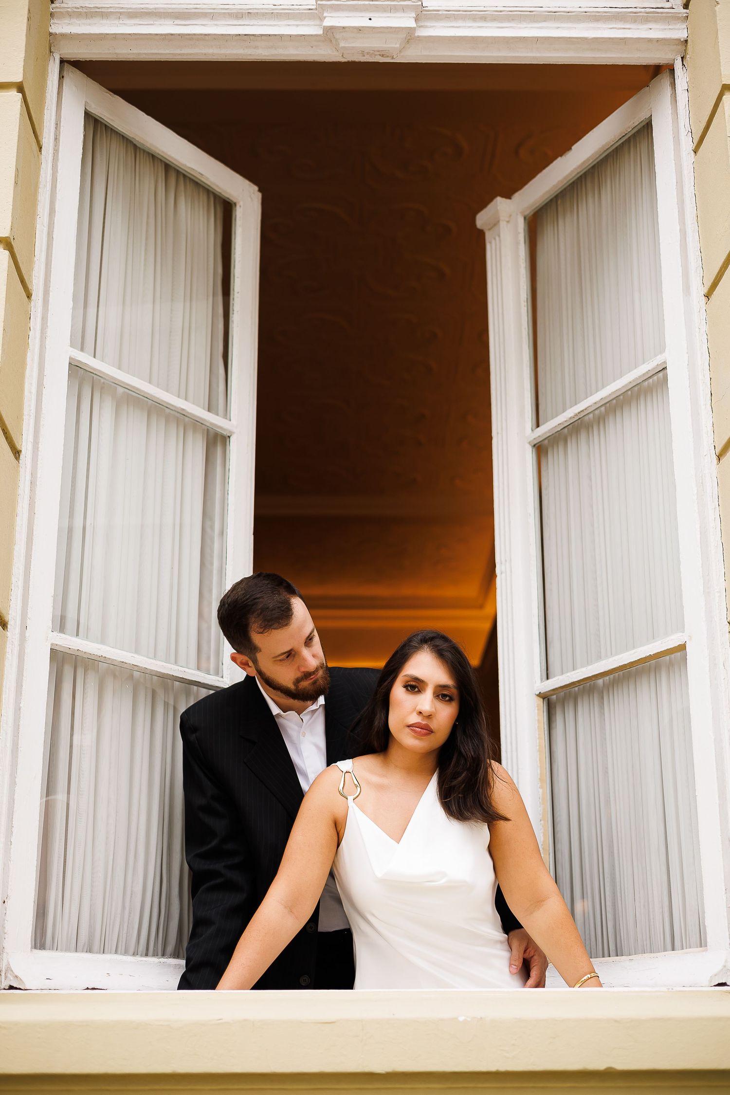 Bride and groom standing in open white French doors at sunset, woman in white dress, man in black suit.