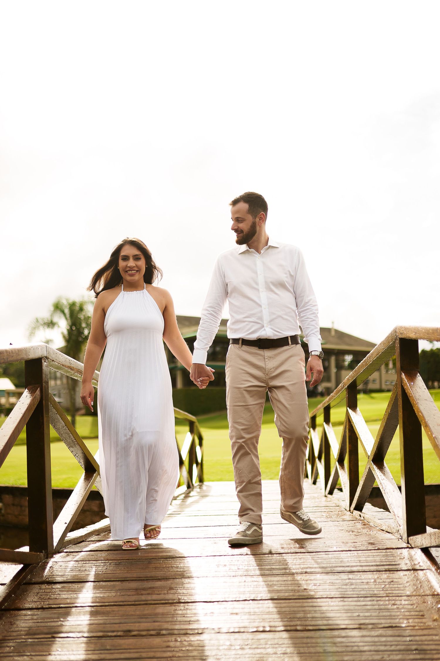 Couple holding hands walking on wooden bridge, woman in white dress, man in white shirt and khaki pants, lush green background.