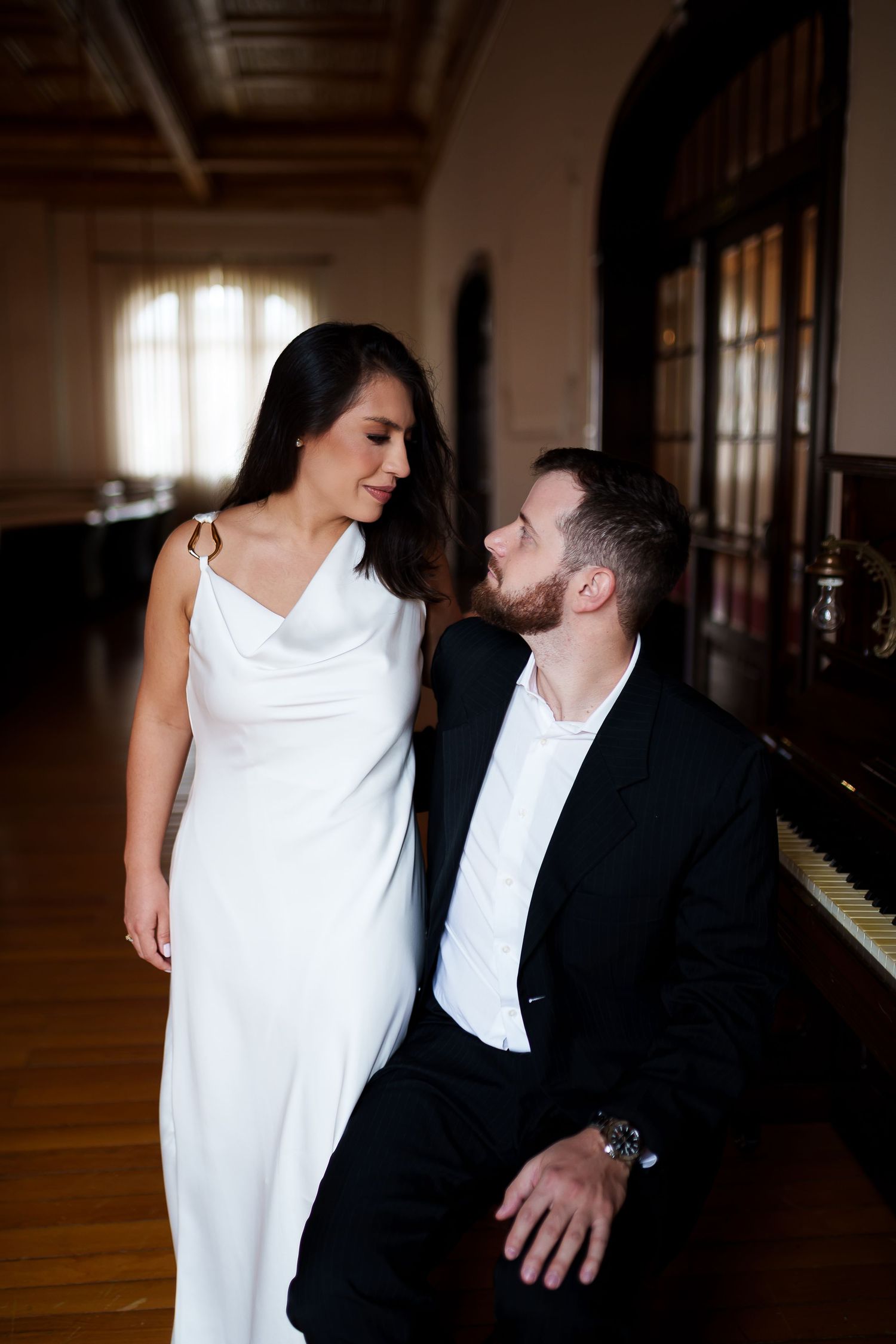 Elegant couple in formal attire sharing a romantic moment near a grand piano in a classic ballroom.