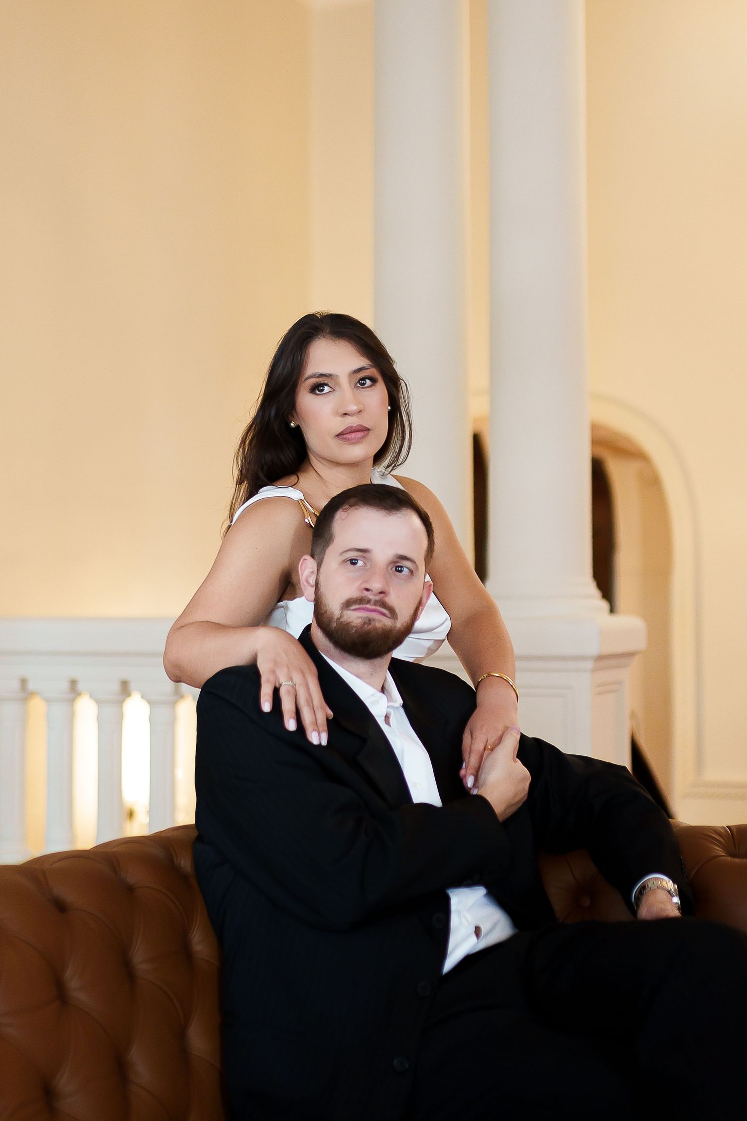 Elegant couple posing in a grand venue, woman in white dress embracing seated man in black suit on leather sofa.