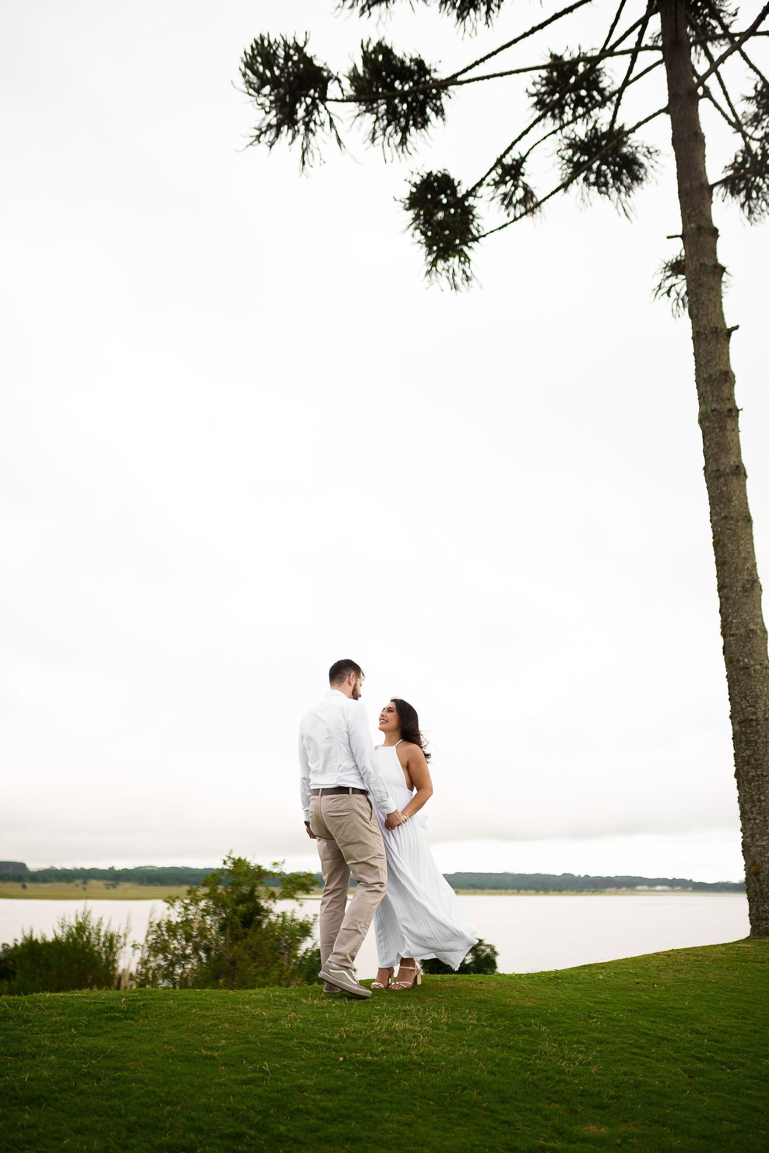 Couple in white attire standing on green lawn near lake with tall pine tree, romantic outdoor portrait.