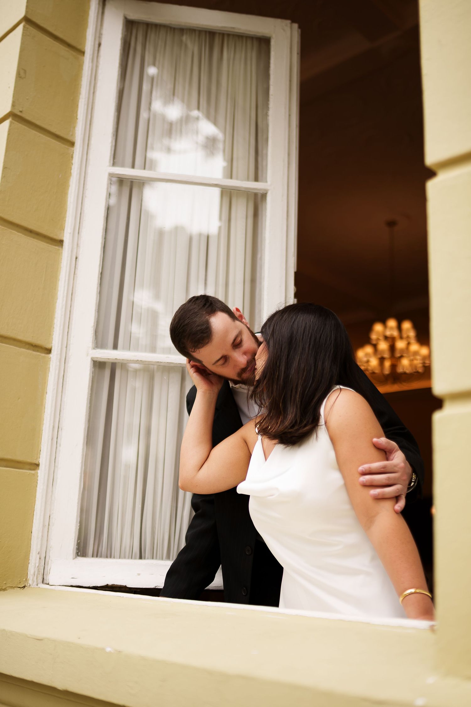 Bride and groom share a romantic kiss by an open window, with a chandelier glowing warmly in the background.