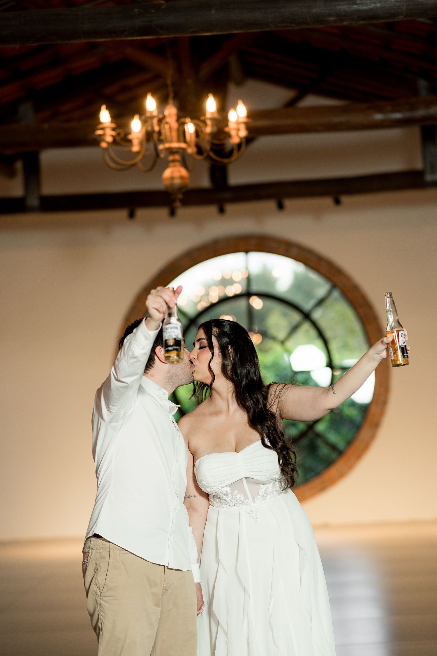 A bride and groom toast with beer bottles under a chandelier in a rustic venue with a round window backdrop.