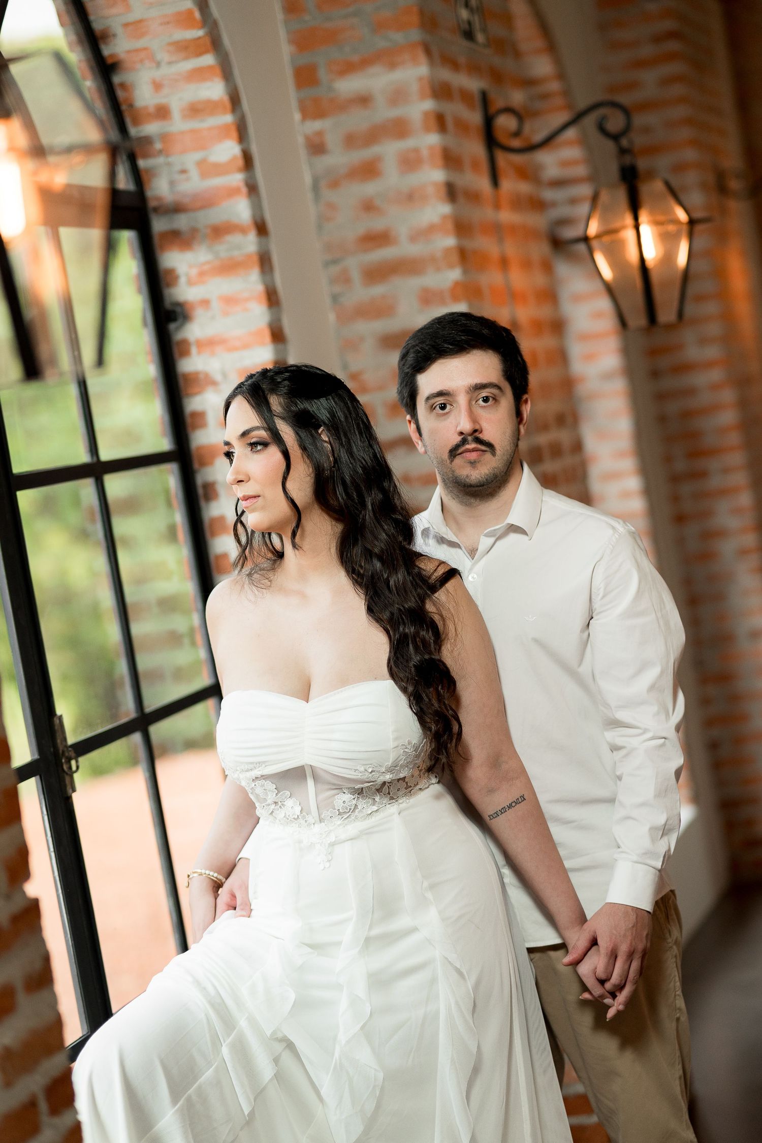 Elegant bride in white strapless gown holds hands with groom in white shirt, posing near brick wall and iron windows.