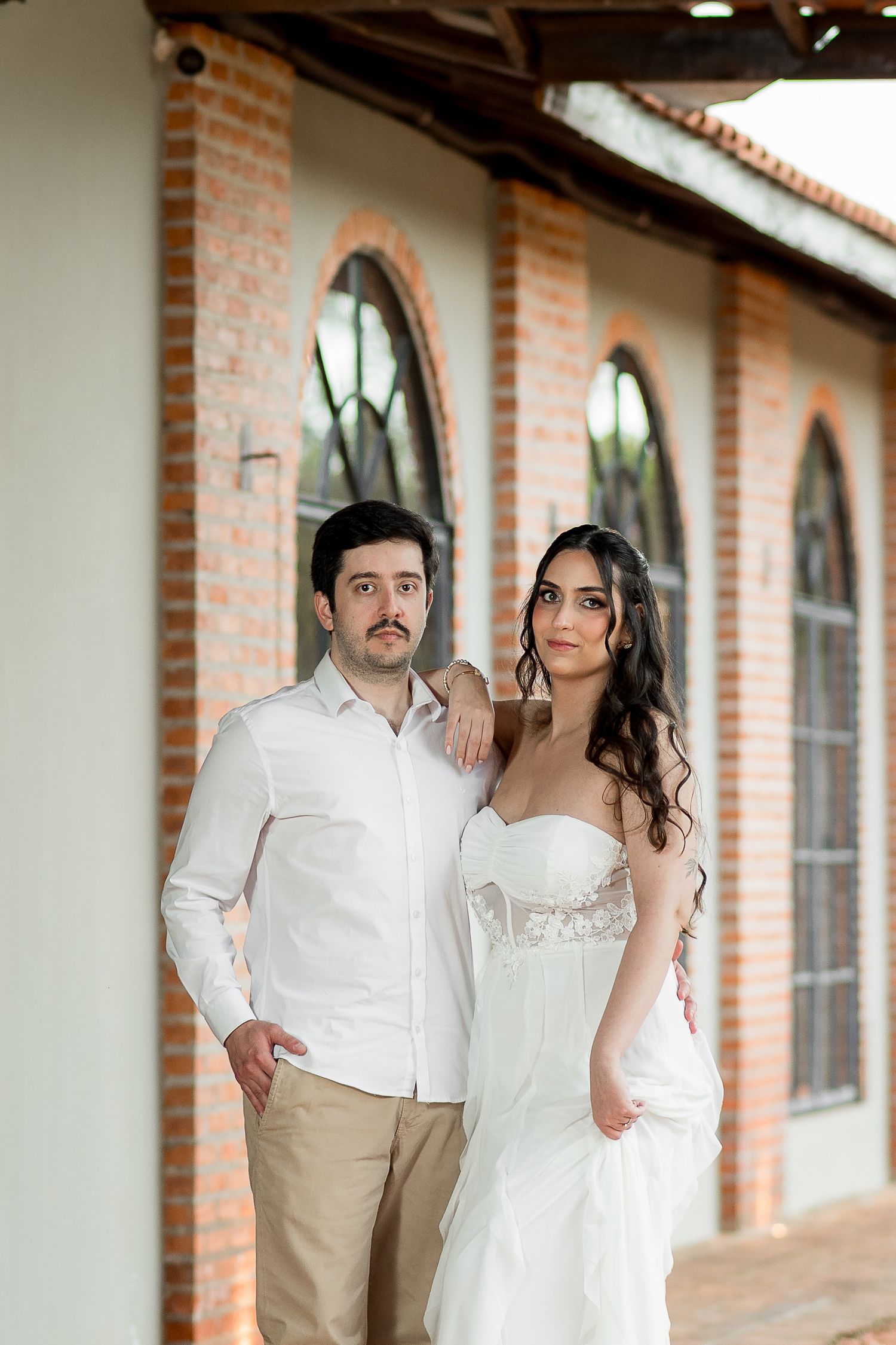 Elegant couple in wedding attire posing before arched windows of a rustic brick building.