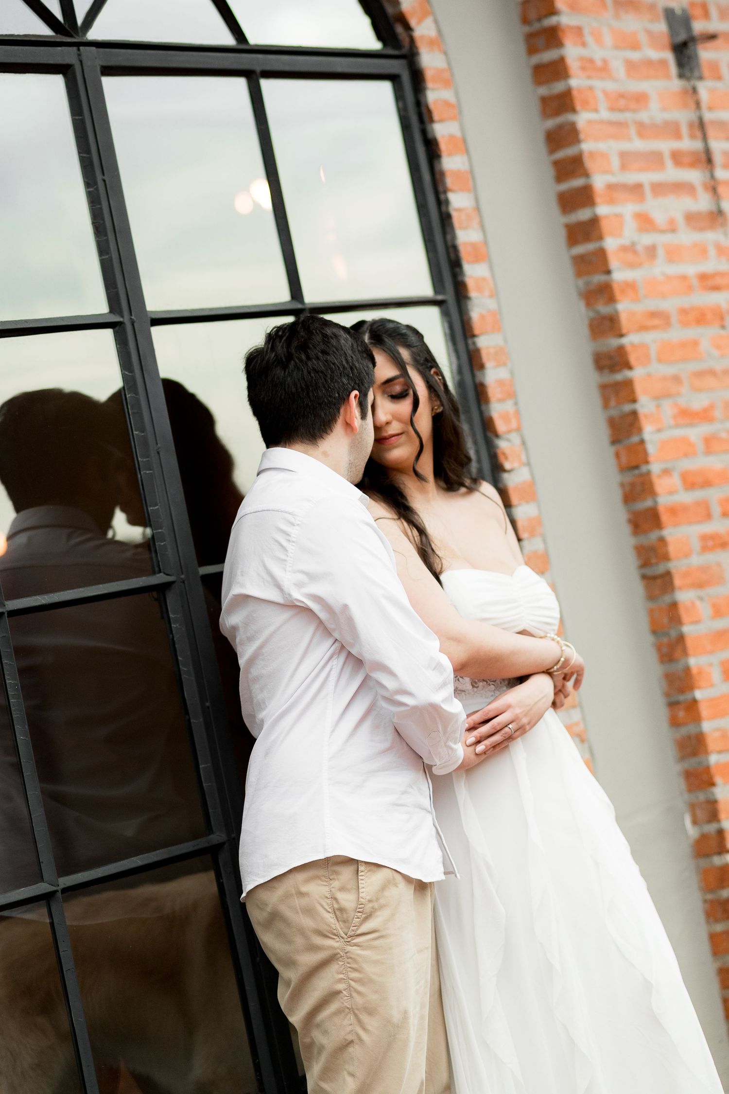 A couple in white wedding attire embraces tenderly near a large black-framed window with brick wall backdrop.