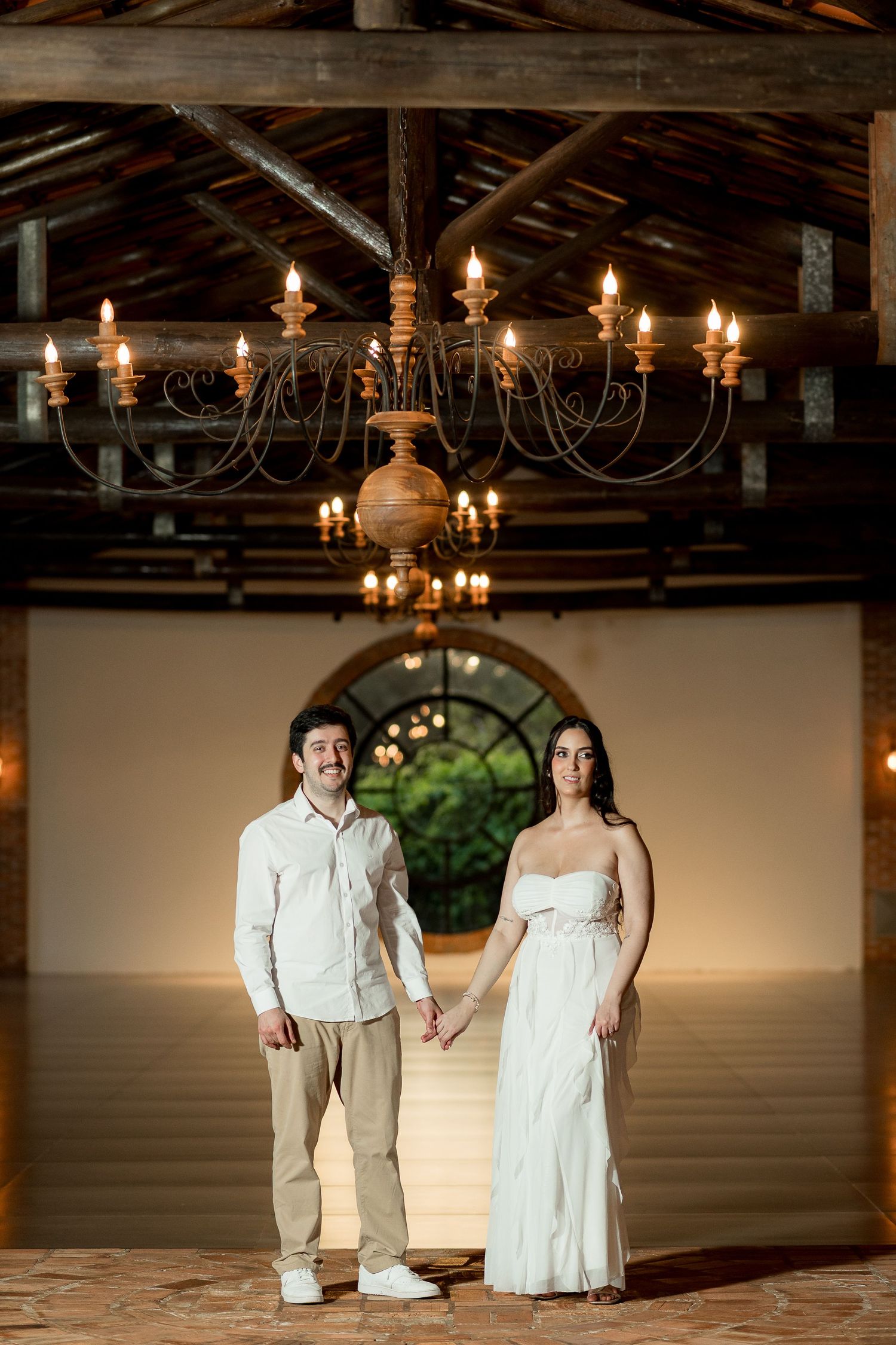 Couple holding hands under rustic chandelier in elegant open-air venue, man in white shirt and woman in white dress.