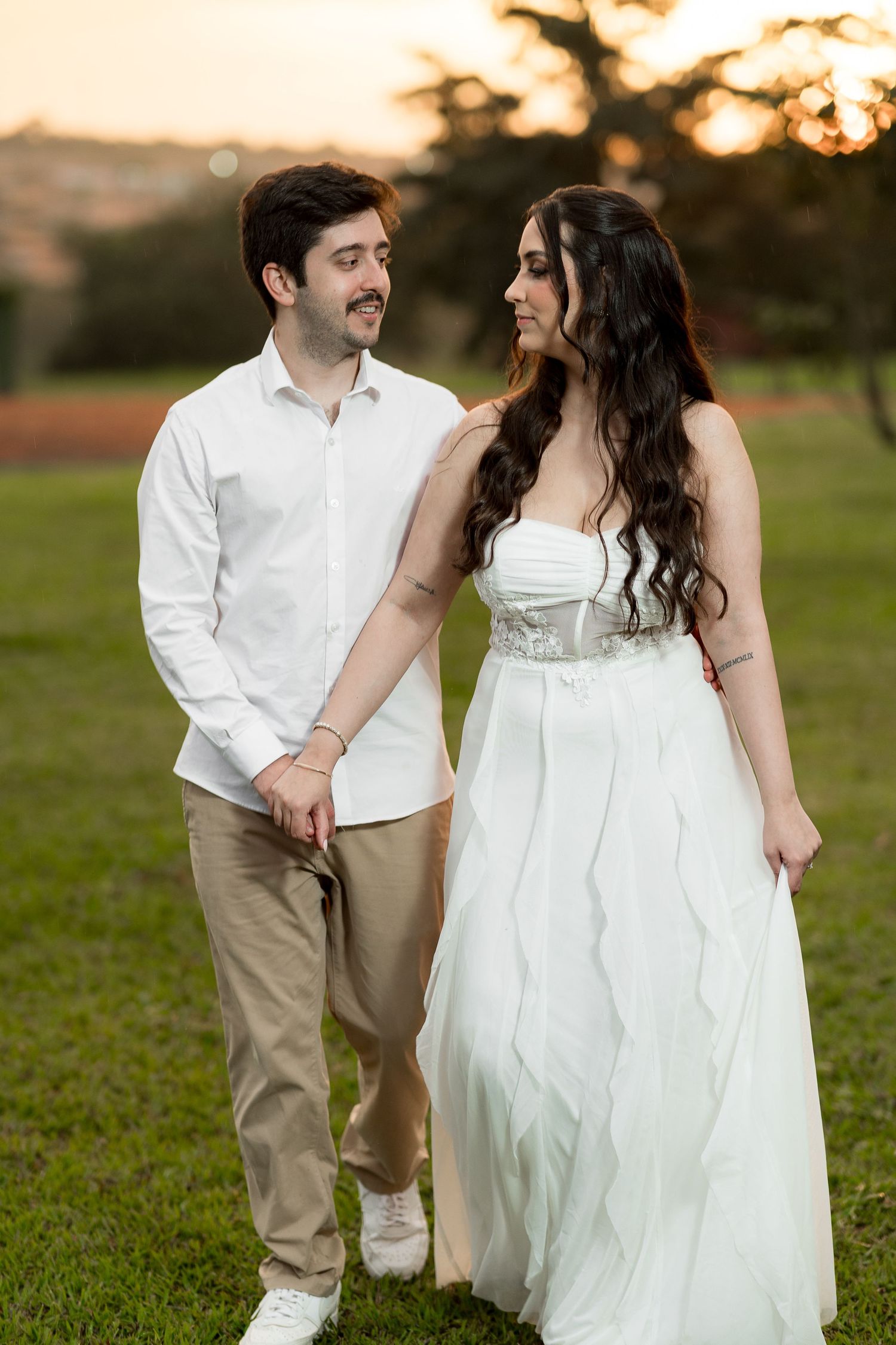 Couple in white attire walking hand in hand at sunset in a lush green park, sharing a romantic glance.