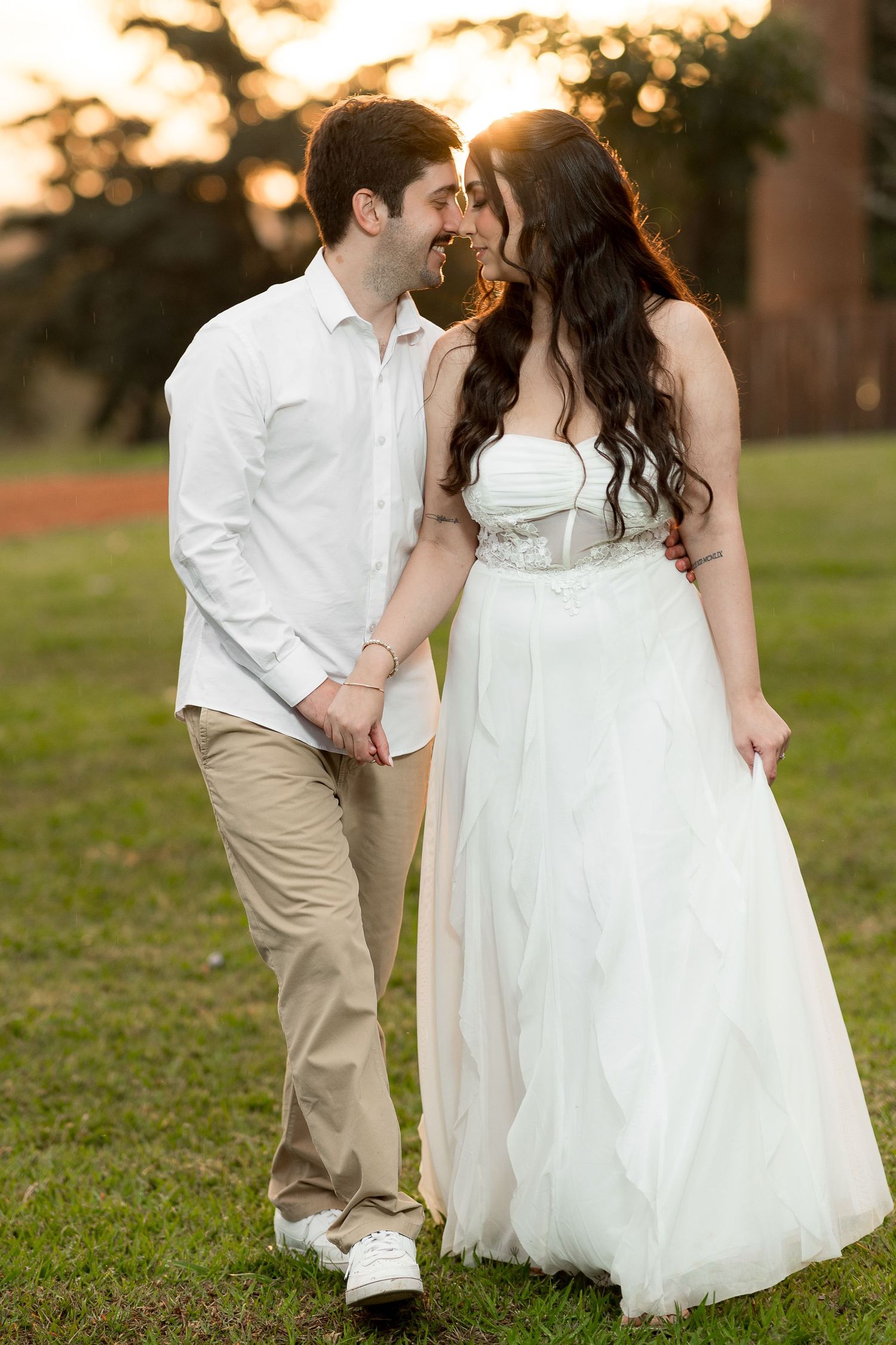 Couple in wedding attire holding hands and sharing a romantic moment outdoors at sunset on green grass.