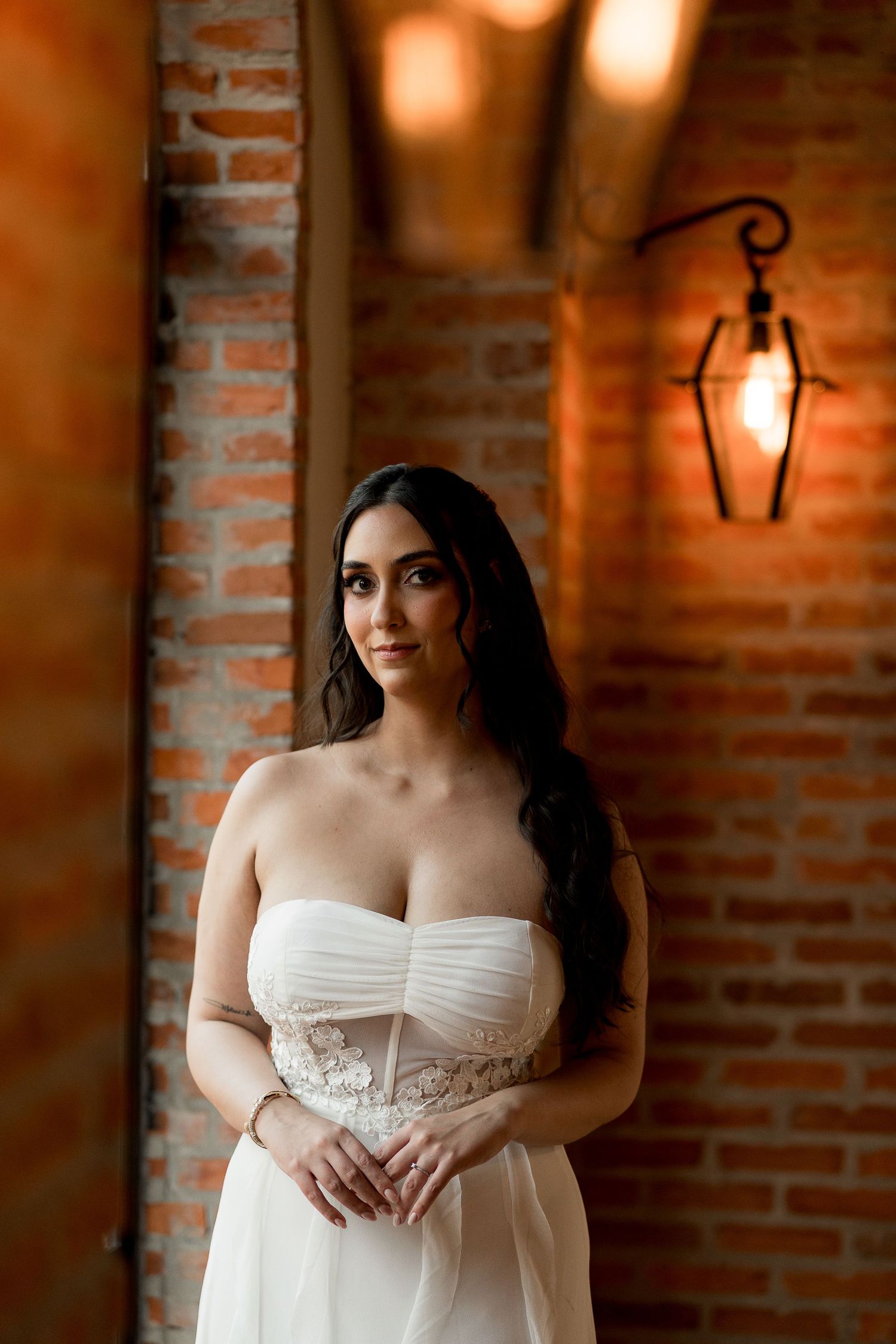 Bride in strapless white lace wedding gown poses near brick wall with vintage lantern in warm romantic light.