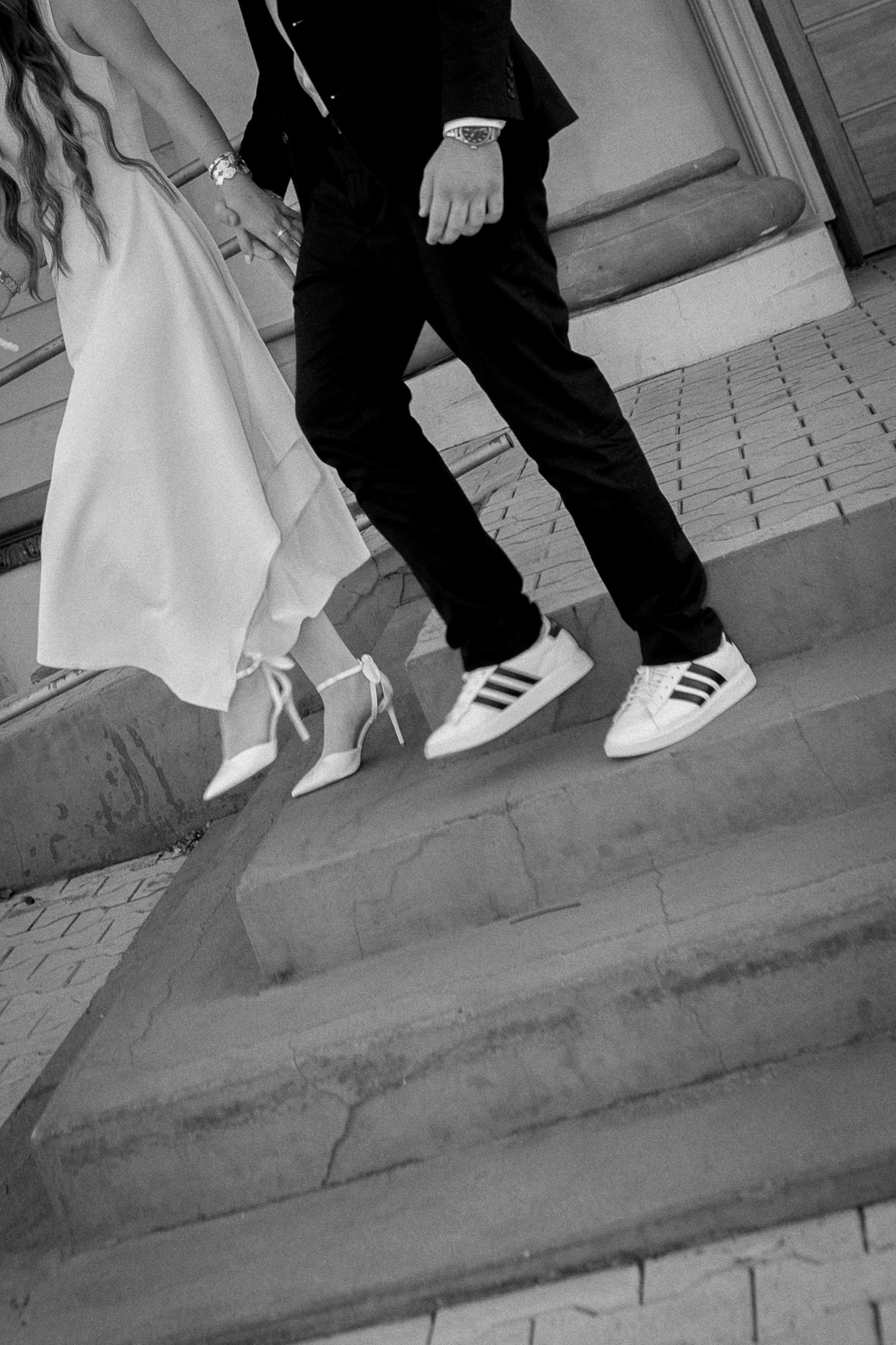 Black and white photo of bride in heels and groom in Adidas sneakers walking down stone steps.