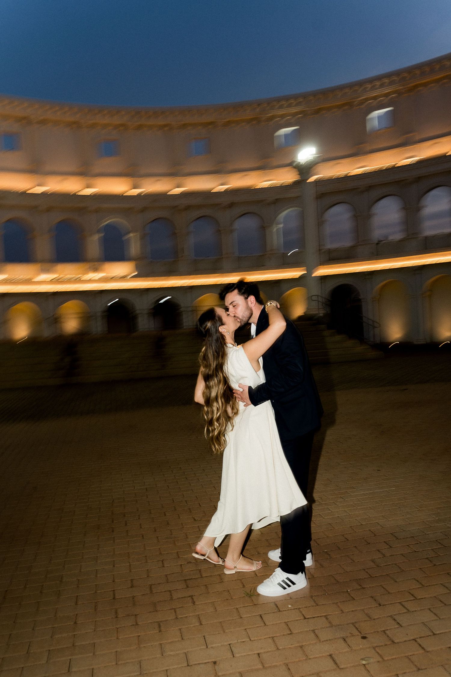 Couple sharing a romantic kiss at night inside an illuminated ancient Roman amphitheater.