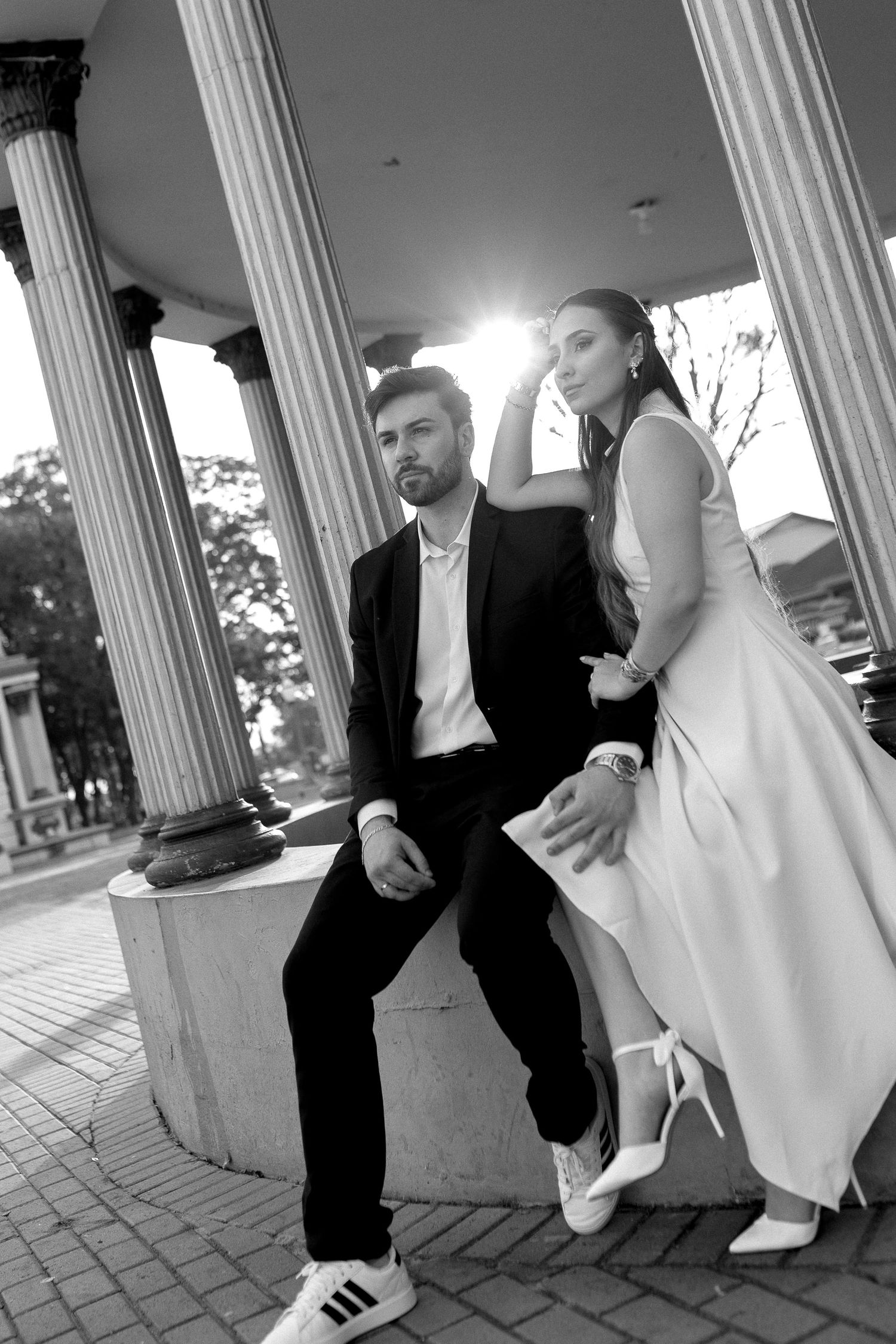 Black and white photo of an elegant couple posing between classical columns, man in suit with sneakers, woman in white dress.