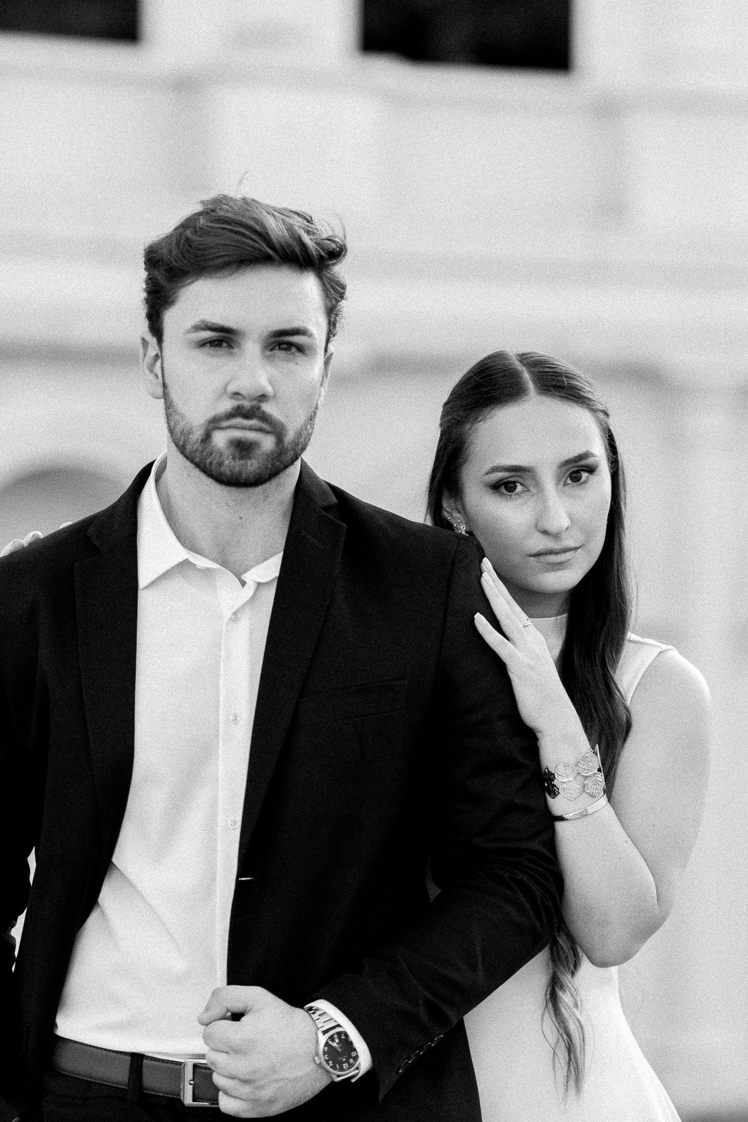 Black and white portrait of an elegant couple in formal attire posing outdoors, man in suit, woman in sleeveless dress.