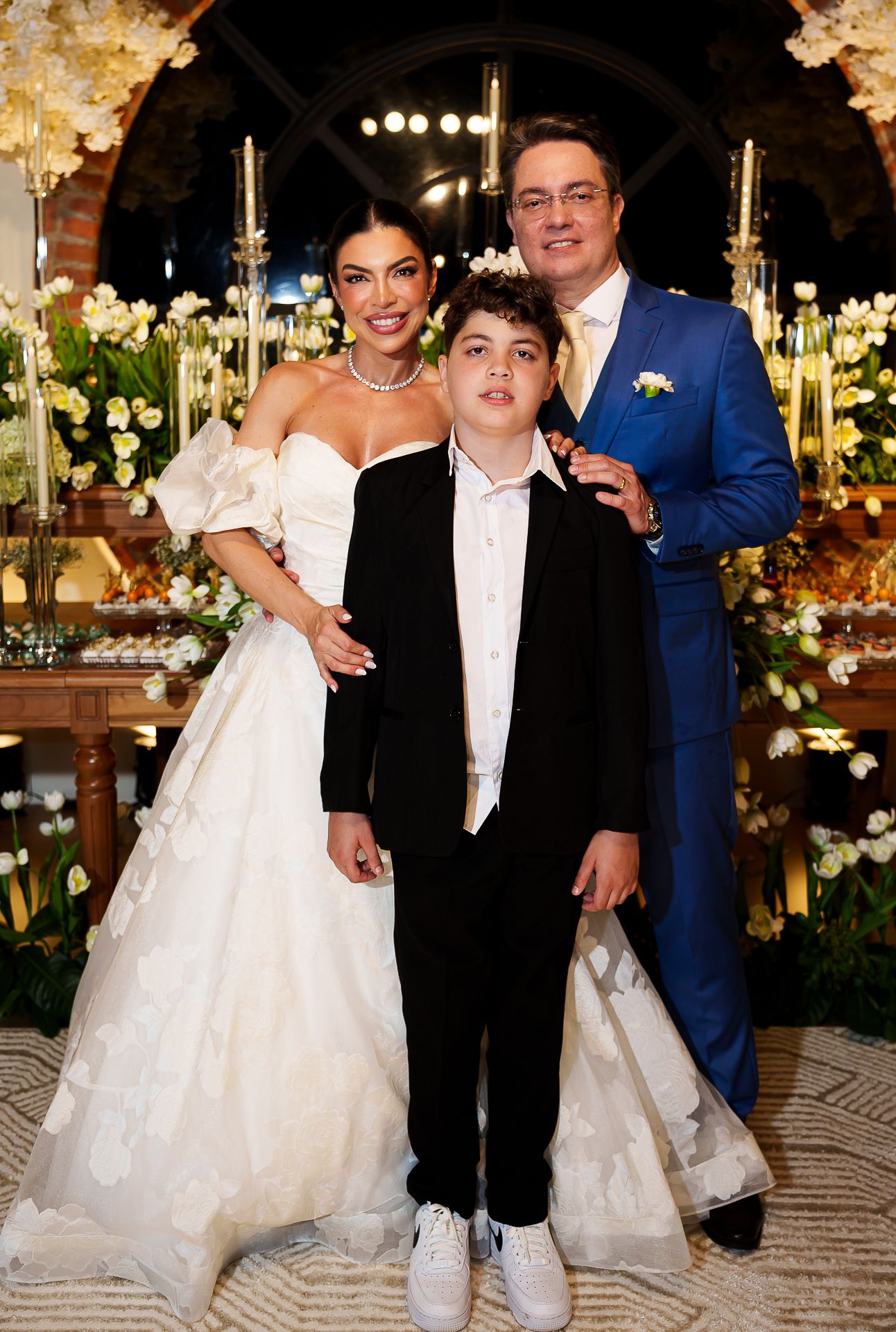 A bride in a white gown, groom in blue suit, and young boy in black tuxedo pose together at an elegant wedding reception.