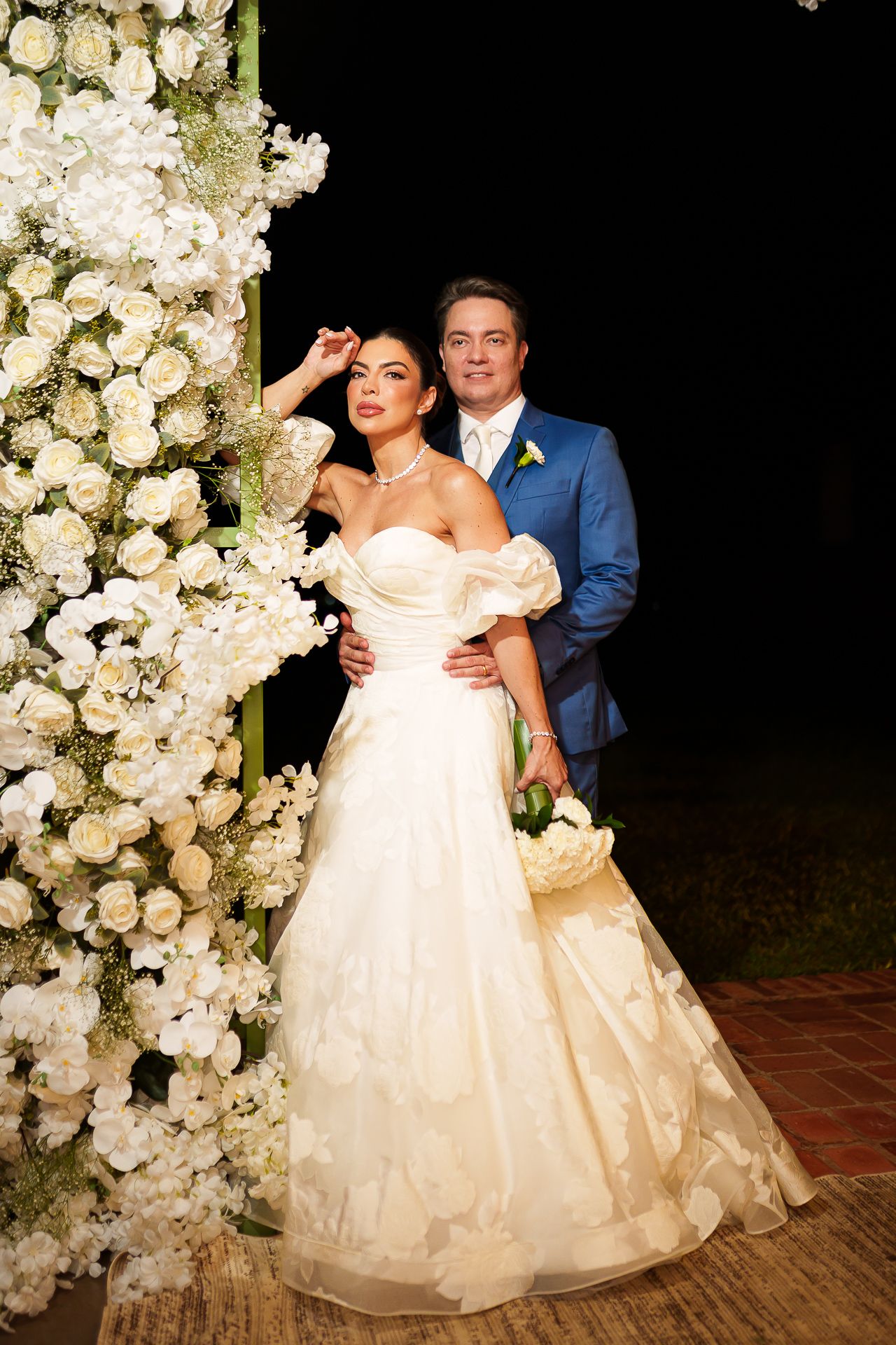Bride in white gown and groom in blue suit pose together by white floral arch at elegant outdoor wedding ceremony.