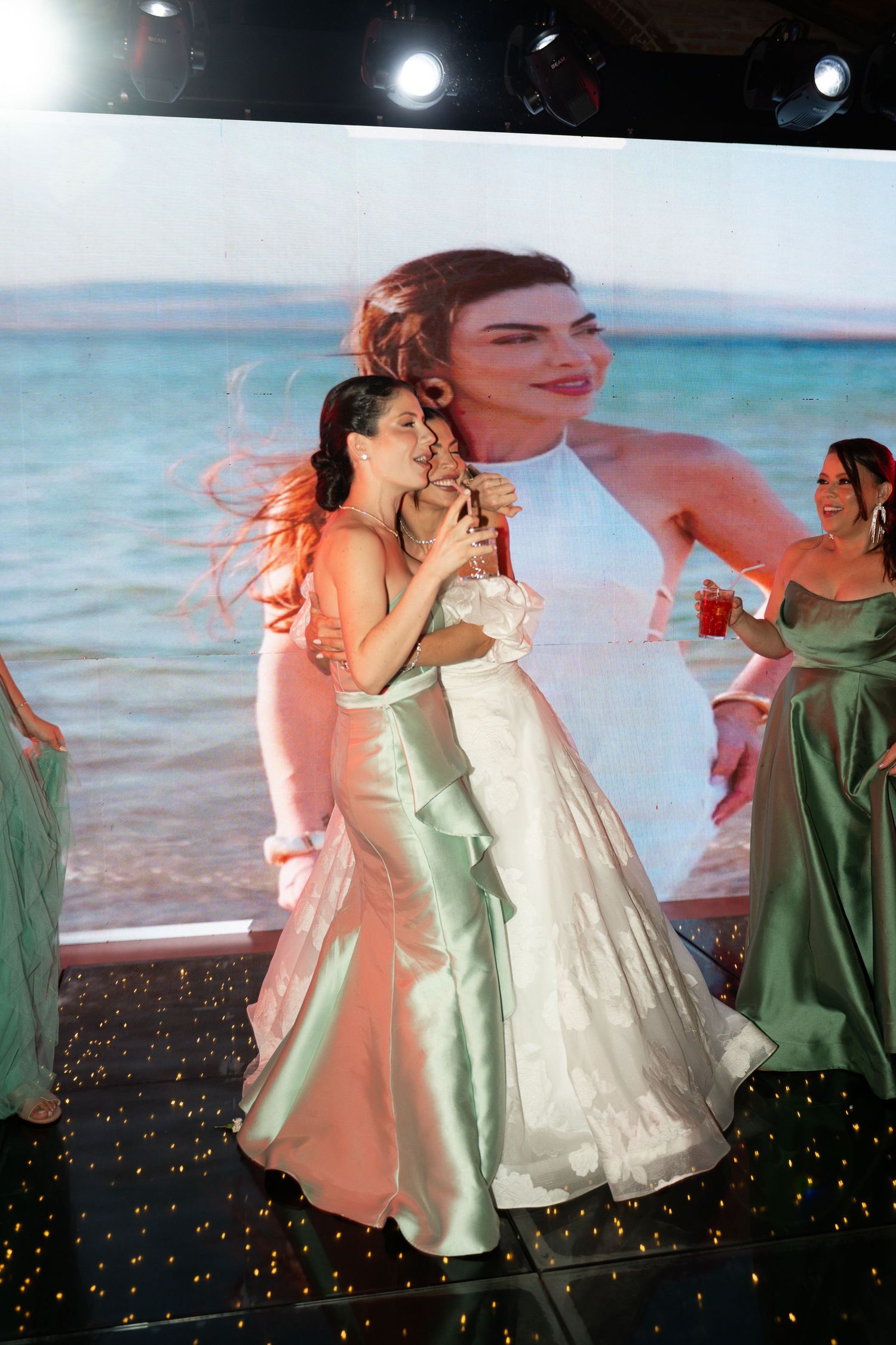 Bride in white gown embraces bridesmaid in mint green dress on dance floor with large screen backdrop.