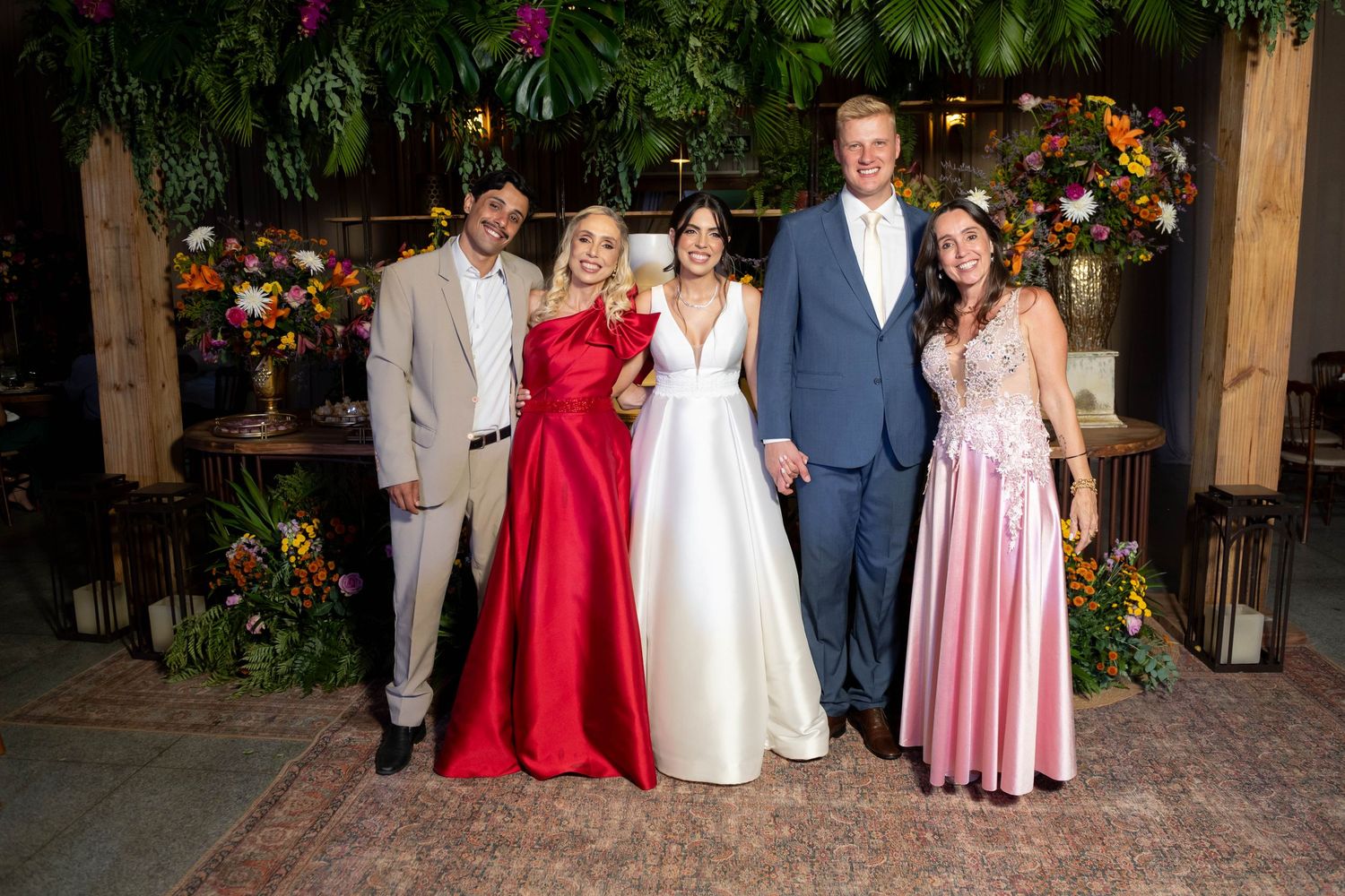 Wedding party posing together, bride in white gown with guests in red, pink, and blue formal attire, floral backdrop.