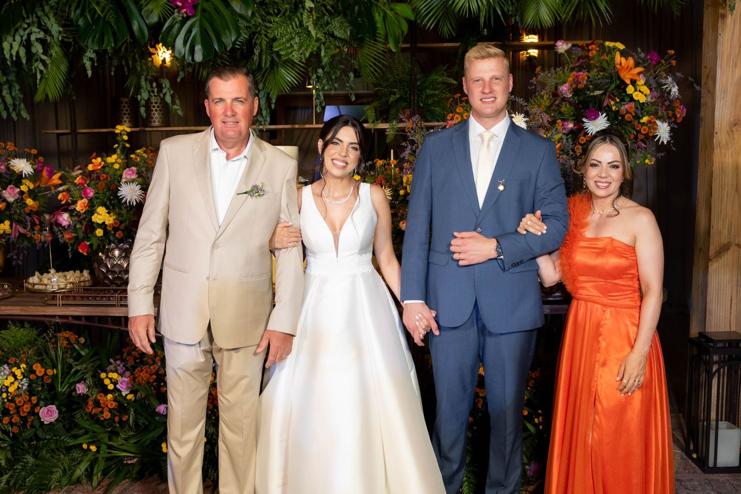 Wedding party of four posing with bride in white gown and guests in formal attire amid lush floral backdrop.