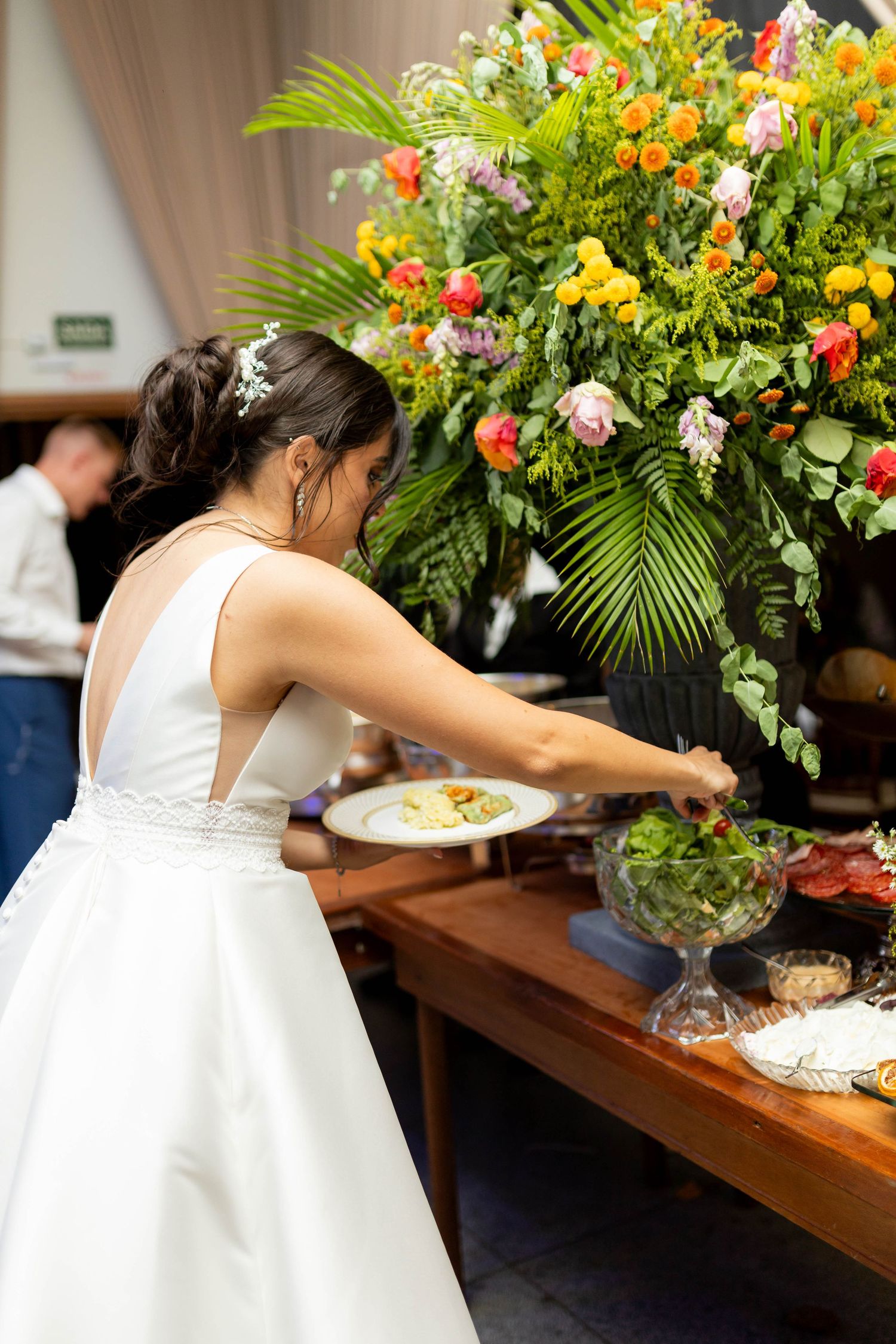 Bride in white gown serving herself at a wedding reception buffet table adorned with colorful floral arrangements.
