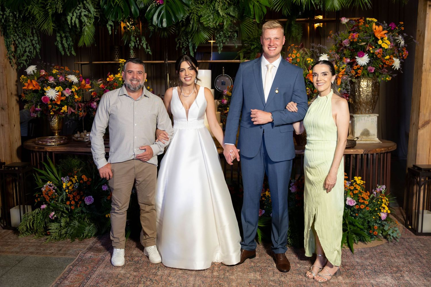 Bride in white gown poses with groom in blue suit and two guests at a floral-decorated wedding reception.